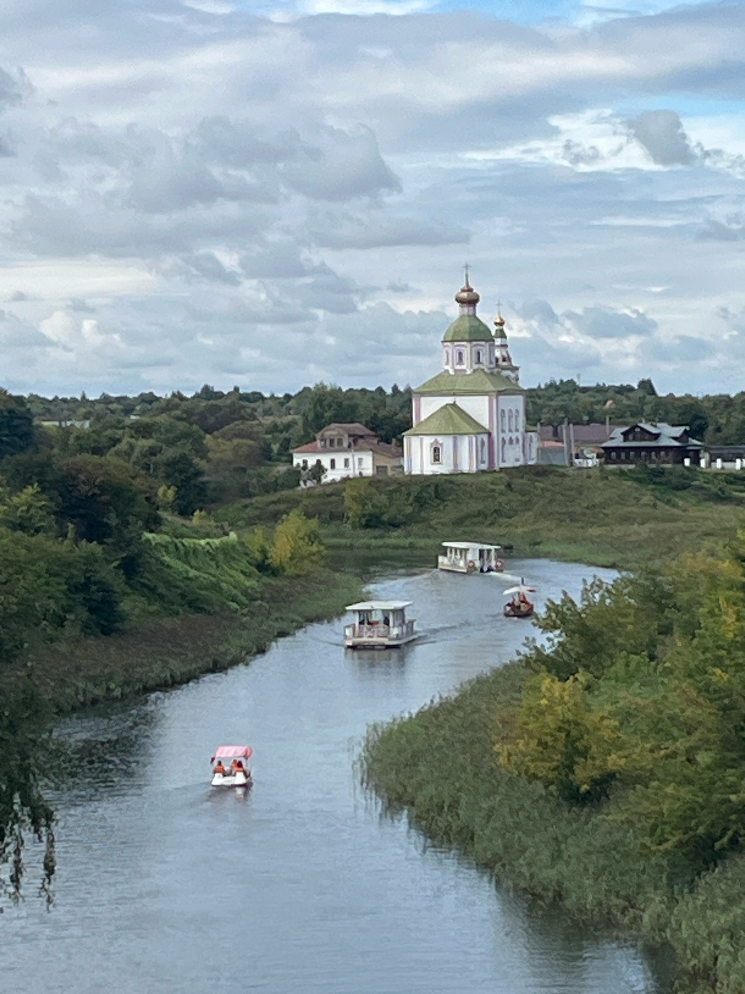 The image depicts a serene river scene with a prominent church in the background, identified as the Church of the Transfiguration in Suzdal, Russia. The church features white walls with pink accents and a green roof topped with golden domes. Several small boats are navigating the river. In the foreground, a white boat with a pink canopy carries two individuals. Further up the river, another white boat with a flat roof is visible, and a third boat with a distinctive sail-like canopy is also present. The riverbanks are lined with dense green foliage, and the background is a rolling landscape of trees and buildings. The sky is overcast with dramatic clouds.