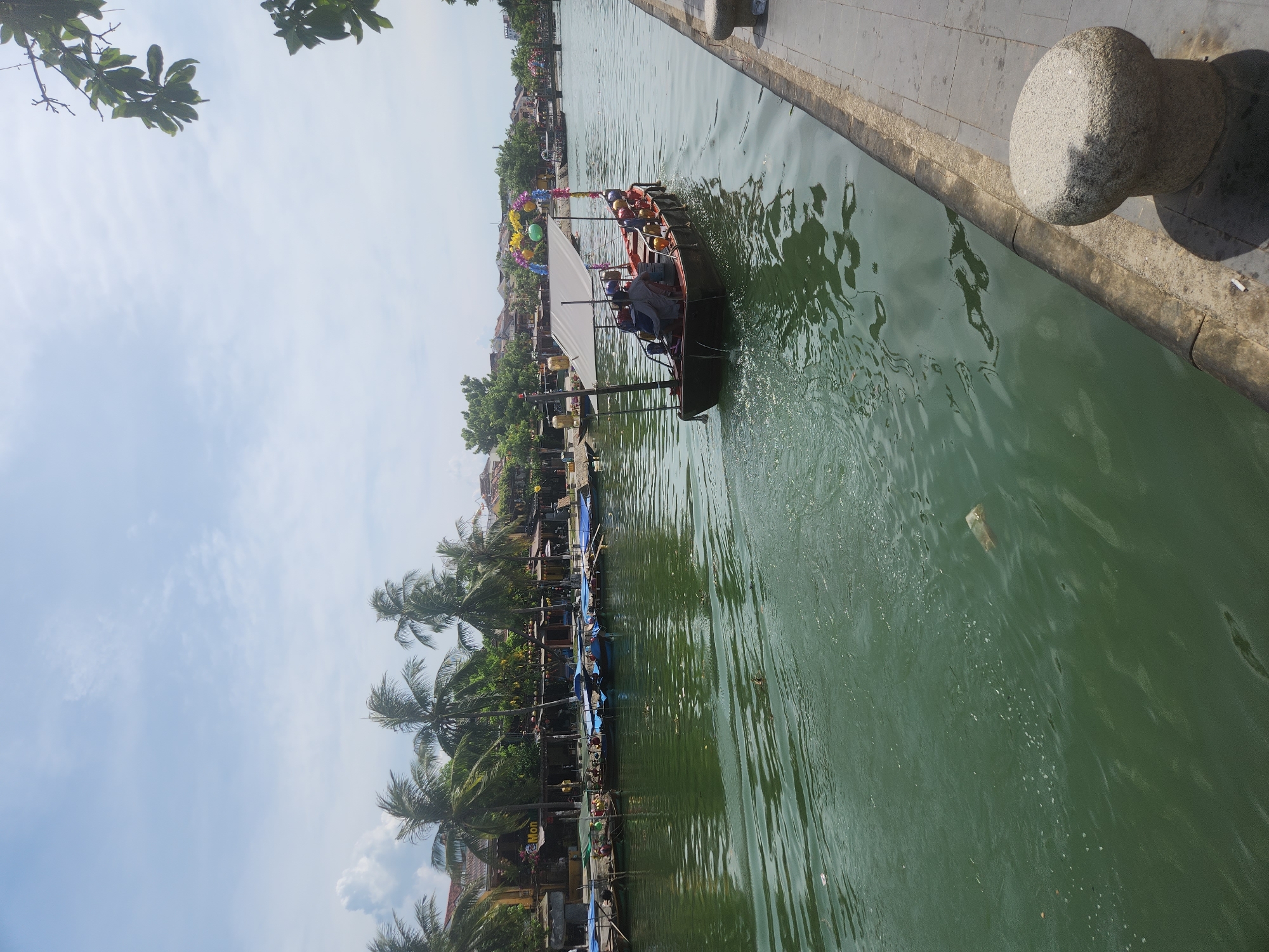 A wide-angle shot captures a canal scene in Hội An, Vietnam. The water is a murky green, with a white plastic bag floating near the left side. On the right bank, a concrete walkway is visible, with a large, rounded stone bollard. A small, dark boat with a white canopy is in the middle of the canal, heading away from the viewer. The boat has a few people on board and is decorated with colorful balloons and streamers. Further down the canal, more boats are docked along the left bank, which is lined with buildings and palm trees. The sky above is a pale blue with scattered clouds.