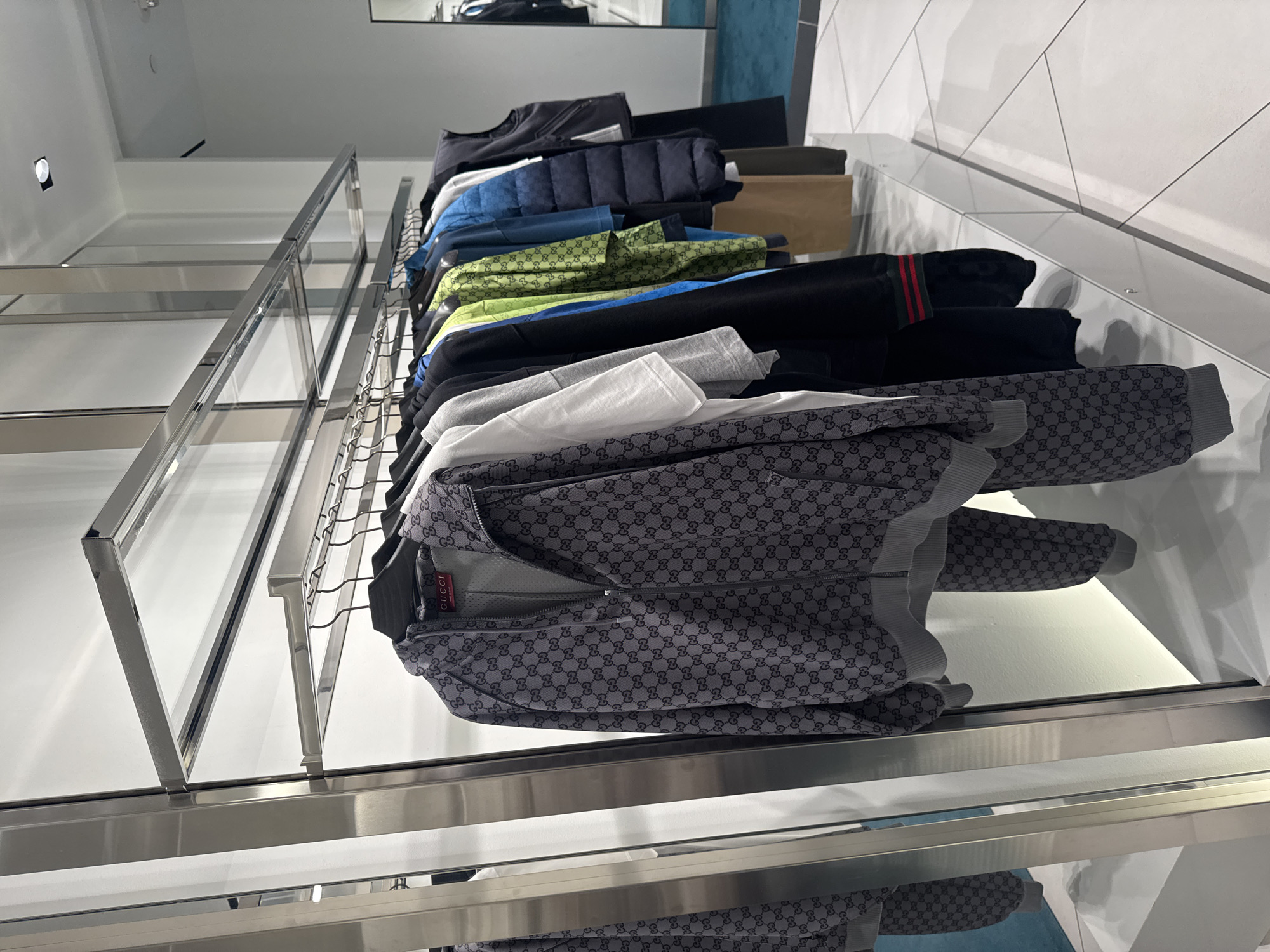 An eye-level shot captures a display of various luxury clothing items arranged on a modern, multi-tiered metal shelving unit within a retail store. The perspective is angled downwards, showcasing the clothes extending from the top right towards the bottom right of the frame, with the sleek metal framework occupying the left side.

No people are visible in the scene. The clothing includes a range of garments, from jackets and hoodies to t-shirts and sweatpants. Notable items include a dark puffer jacket, a blue top, a bright lime green patterned jacket, and several black items, one with distinctive red and green stripes on a cuff. A stack of white and grey t-shirts is also present.

The most prominent items in the foreground are a grey full-zip hoodie and matching sweatpants, both featuring the iconic "GG" monogram pattern from Gucci. A red label with the brand name "GUCCI" printed in white text is clearly visible on the hoodie. Another item, likely a green jacket, also shows a repeated "GG" logo pattern.

The background consists of a clean white wall with subtle diagonal paneling. A reflective surface, possibly a polished floor or lower shelving, mirrors the clothing display at the bottom of the image. The setting suggests a well-maintained, high-end retail environment, likely a Gucci store in Rosemont, United States of America, dedicated to showcasing its apparel. The lighting is bright and even, highlighting the products.