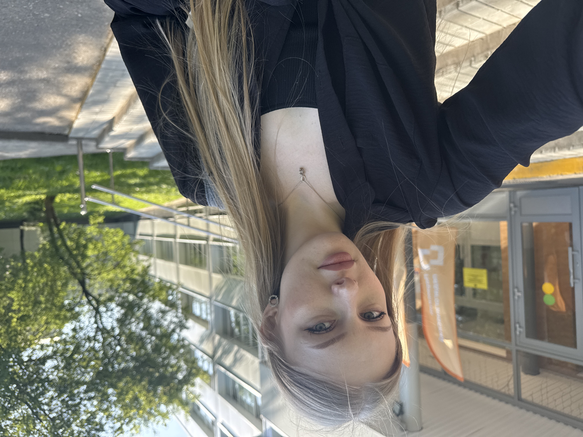 A young woman with long blonde hair and fair skin poses for a selfie outdoors. She is wearing a black top and a black jacket, and a delicate silver necklace. The background includes a modern building with glass windows and a silver metal frame. To the left, there are green trees and bushes. A portion of a walkway or patio made of light-colored stone is also visible. Given the signage and architecture, the location is most likely in Perm, Russia. The photograph appears to be taken during the day, with sunlight illuminating the scene.