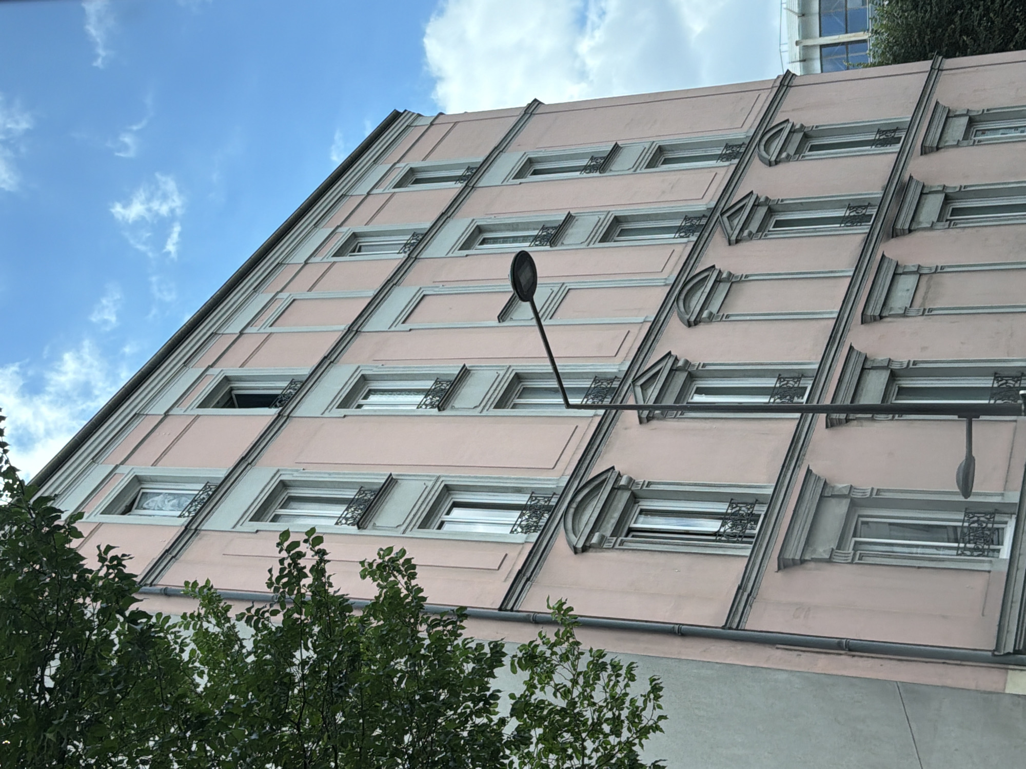 The media file depicts a close-up, low-angle view of a pink apartment building with a pale gray trim, seen against a blue sky with scattered white clouds. The building has a series of evenly spaced rectangular windows, many of which are adorned with decorative black wrought-iron grilles. The architecture features repeating geometric patterns and layered details along its facade. In the foreground, the tops of lush green trees obscure the lower portion of the building. A black streetlamp is visible extending from the building's facade, adding a functional element to the scene. The lighting suggests it is daytime, with no specific indication of time beyond that. The location context is Ivry-sur-Seine, France. There is no discernible activity or event taking place. No text is visible in the image.