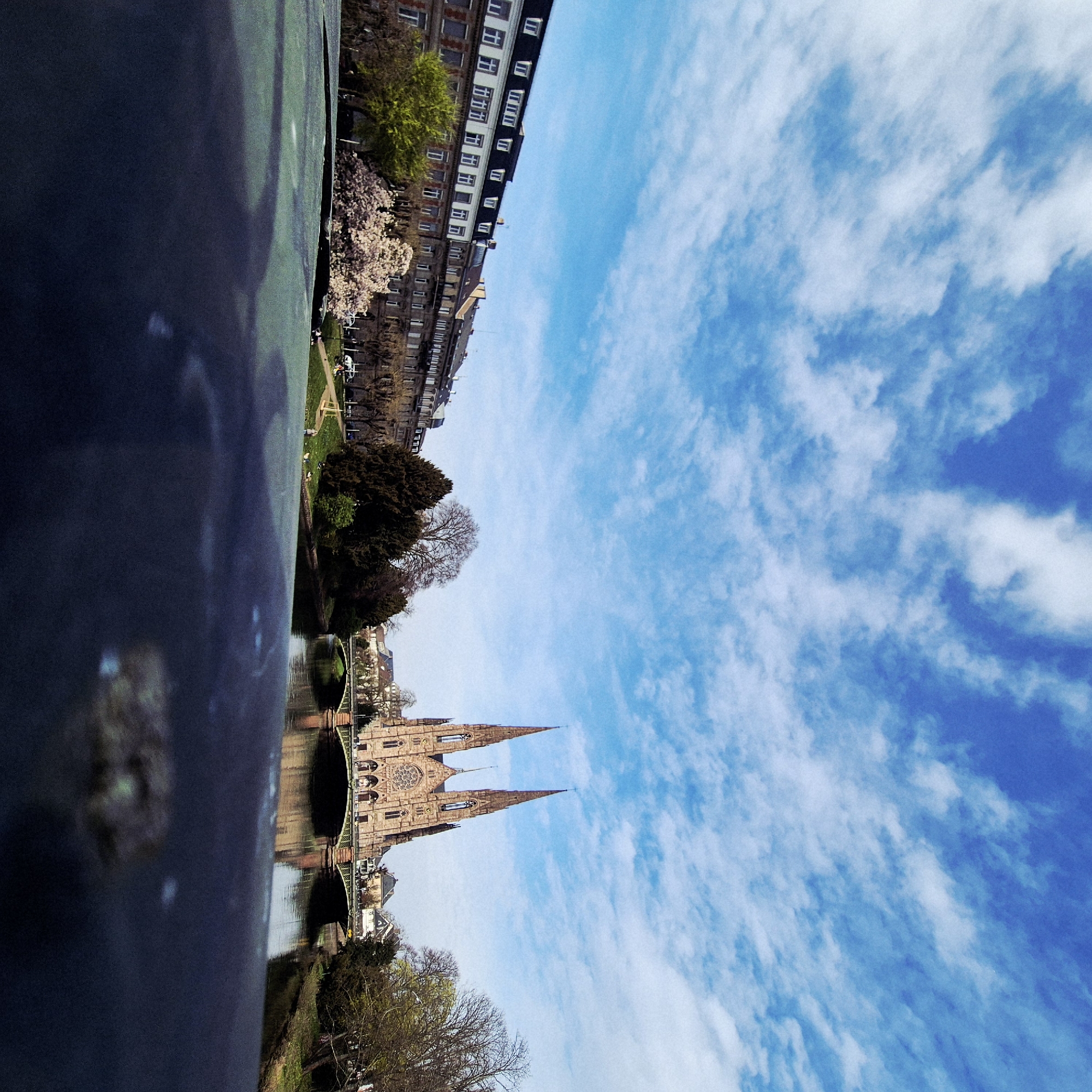 The image presents a wide-angle, slightly low-angle view of the Église Saint-Paul and its surrounding environment in Strasbourg, France. The viewpoint appears to be from a bridge or elevated structure, with a dark, textured, out-of-focus surface occupying the lower foreground, possibly a bridge railing.
The scene is dominated by the Neo-Gothic Église Saint-Paul, centrally located in the midground, featuring two prominent, ornate spires and a large rose window on its reddish-brown stone facade. It is situated on an island, flanked by a calm body of water, identified as the Ill River, which reflects the sky and architectural elements. A multi-arch bridge, likely the Pont Saint-Paul, with decorative green railings, spans the river directly in front of the church, connecting the banks.
On the left bank of the river, a continuous row of multi-story European-style buildings is visible, characterized by dark sloping roofs with white dormer windows, numerous windows along their facades, and some balconies. The ground beneath these buildings features a paved area (road or sidewalk) and a grassy bank. Trees with varying foliage, including one with bright green leaves and another displaying light pink blossoms, are situated along this bank. Several individuals are present, some seated on the grassy bank, others walking, and at least two white vehicles are visible on the paved area.
The right bank, extending from behind the church, displays more trees, some still bare and others with nascent spring leaves, lining the river. Additional residential-style buildings are discernible behind the church and further to the right. The sky overhead is bright blue with scattered, wispy white clouds. The overall lighting suggests daylight conditions. No overt interactions or actions are clearly discernible beyond pedestrian movement and stationary vehicles.