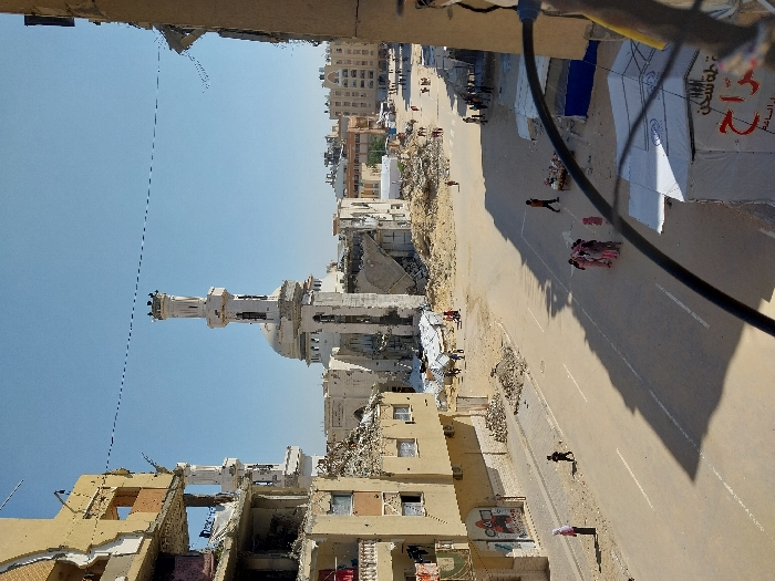 The image depicts a devastated urban street scene in Khan Yunis, Palestinian Territory, under a clear blue sky. In the center-background, the Great Mosque of Khan Yunis is significantly damaged, with its white minaret partially intact but large sections of the dome and surrounding structure reduced to rubble.
To the left, a multi-story residential building shows extensive damage, with exposed interior rooms, broken walls, and debris. Clothes hang from remnants of balconies. Piles of rubble and dirt are scattered along the asphalt road and in open areas where buildings once stood.
Approximately 15-20 individuals are visible throughout the scene. On the left foreground, a person in a pink hijab stands on the sidewalk near a child. A group of 5-6 individuals, including children, are clustered in the middle of the street, while another person walks in the mid-ground. Further back, numerous smaller figures are seen moving amidst the debris. On the right, two white temporary tents with Arabic script (indicating aid) are set up, next to a blue structure and a person pushing a small cart. Long shadows are cast across the street, suggesting bright daylight. A thin wire spans the upper portion of the scene, and a thick black cable is visible in the lower right foreground.