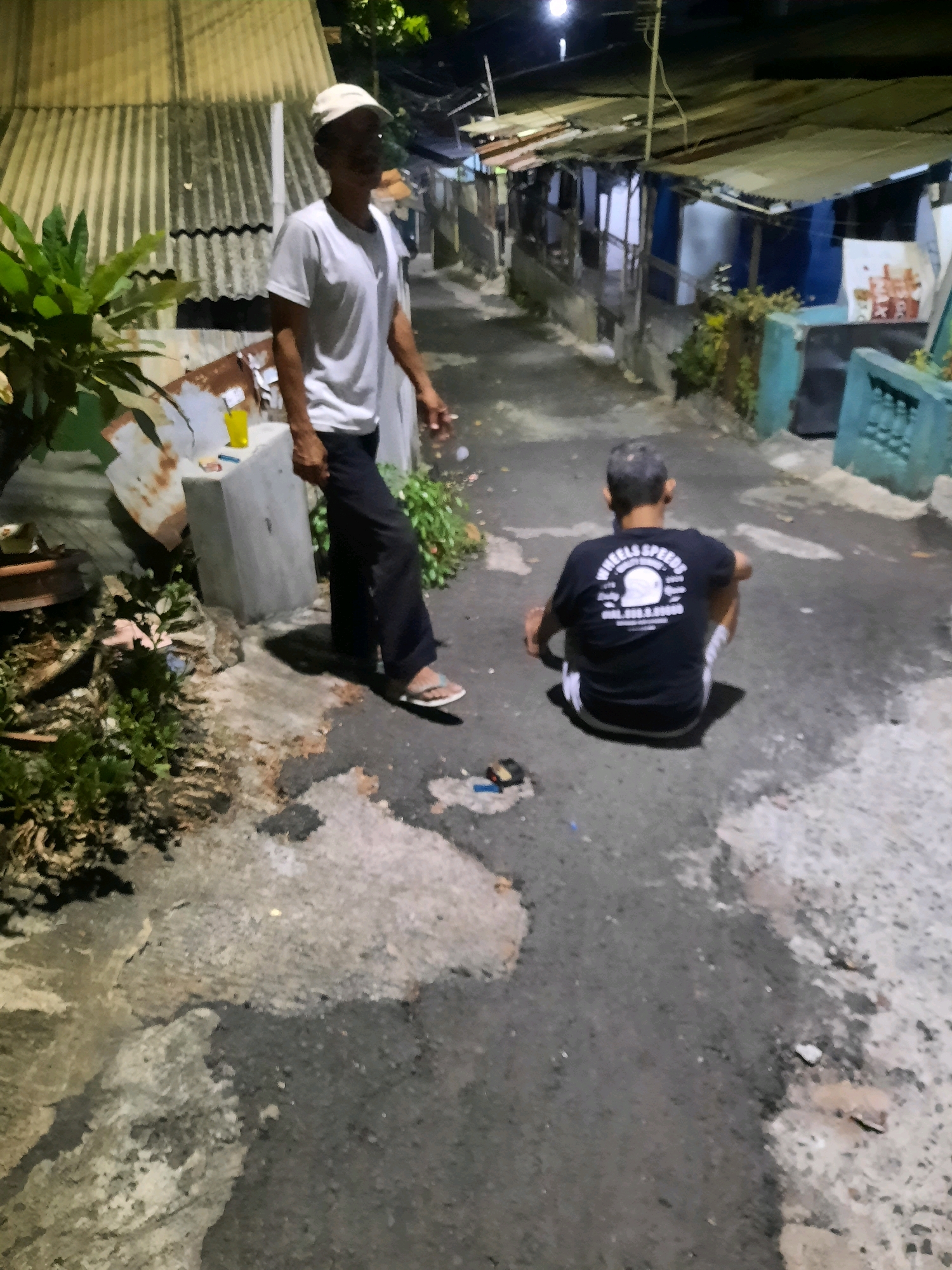 In an alleyway in East Jakarta, Indonesia, at night, two men are visible. One man stands facing to the left, wearing a white t-shirt, dark pants, and a light-colored cap. He has a cigarette in his right hand and appears to be in motion. He is positioned in the foreground, to the left of the frame.
The second man is seated on the ground in the middle of the alleyway, facing away from the camera and towards the right. He is wearing a black t-shirt with a graphic design on the back, dark shorts with white stripes, and appears to have grey hair. He is crouched with his legs folded beneath him.
The environment is an urban alley with corrugated metal roofs and makeshift structures lining the sides. The ground is paved with asphalt and some concrete. There are plants visible on the left side of the frame, and discarded items or debris are scattered on the ground. The lighting suggests it is nighttime, with overhead lights illuminating parts of the scene.