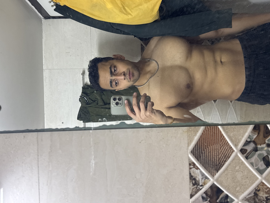 A muscular man poses in a bathroom mirror. He wears dark shorts and a silver chain. His torso is bare, revealing defined chest and abdominal muscles. The mirror reflects a cluttered bathroom. Clothes hang on a rack, and a towel is visible. The bathroom tiles feature a geometric pattern. The man holds a smartphone in his hand, seemingly taking a selfie.