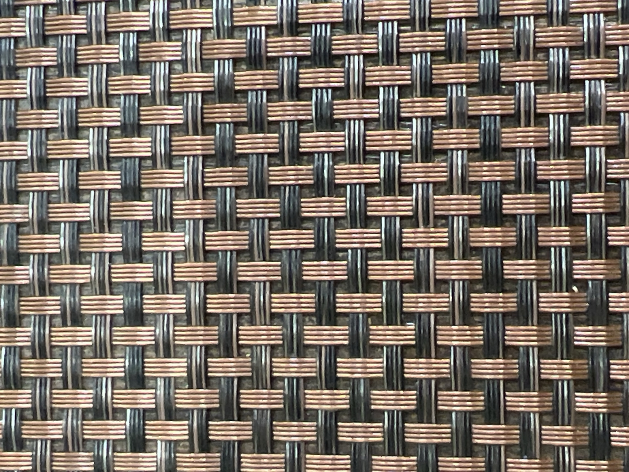 The image shows a close-up, overhead view of a woven mat. The mat is composed of alternating strands of dark grey and copper-colored material, woven together in a simple over-under pattern. The strands are thin and appear to be made of a rigid material, possibly plastic or metal. The lighting suggests the mat is outdoors, as evidenced by the diffused light and slight shadows. There is no indication of the city of Sinyavino, Russia, or any people, actions, or interactions.