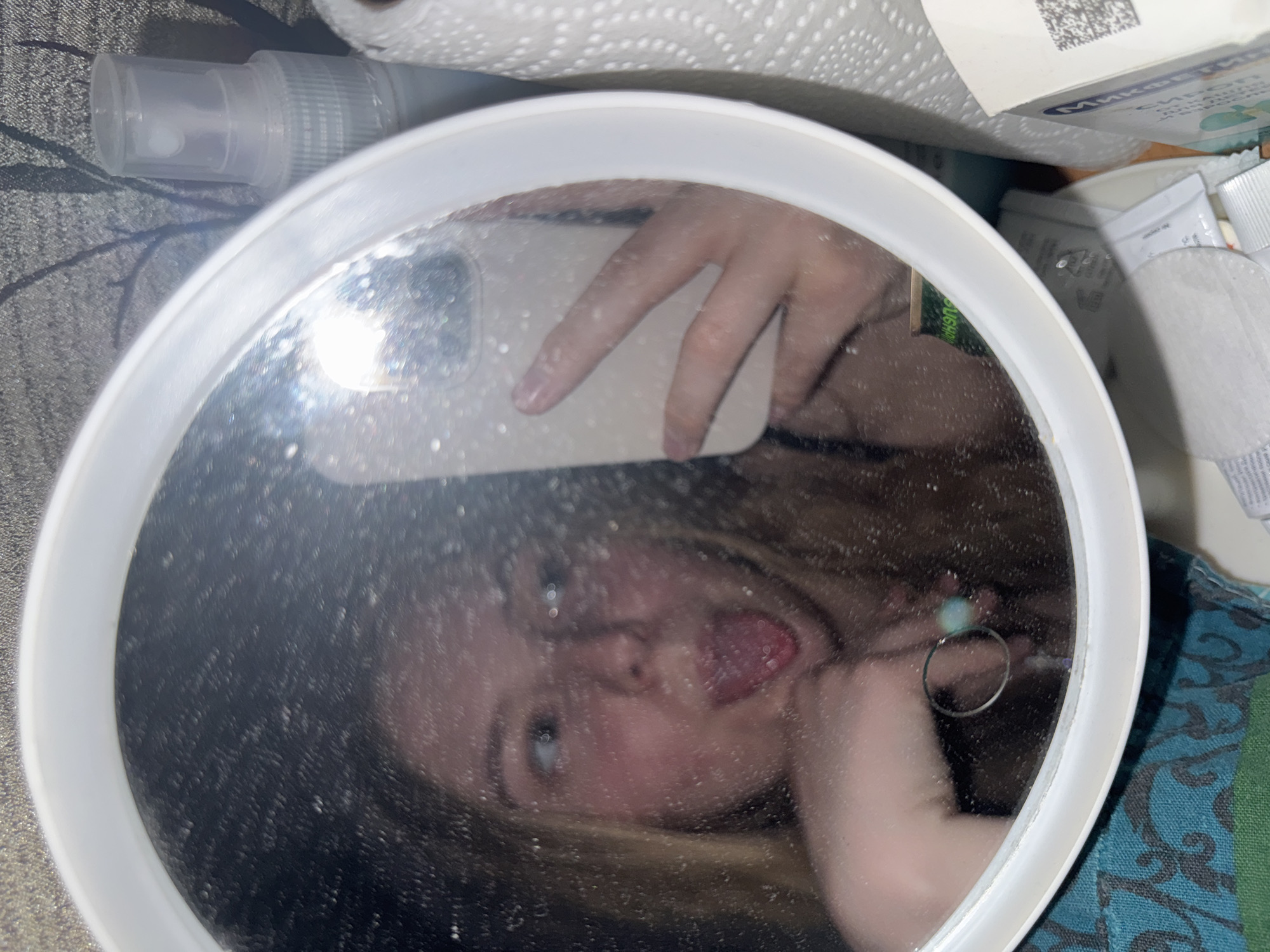 The image depicts a close-up, reflected view of a young female taking a selfie in a round, white-framed mirror in Perm, Russia. The mirror's surface is speckled with numerous small particles or water droplets, causing a diffused and slightly blurry appearance of the reflection. The female has long, light-colored hair and is making a playful expression, sticking her tongue out and looking to her left. Her left hand, visible in the reflection, is positioned under her chin and features a ring with a light blue or green stone. Her right hand is holding a light-colored smartphone, used to capture the image, which is also reflected in the mirror.
The surrounding environment includes a clear plastic spray bottle with a white pump mechanism and a roll of white paper towels situated to the right of the mirror. Below the mirror, a cardboard box with Cyrillic text, including "Мимивелл" and a QR code, is visible. Various small personal care items, such as cotton pads and sachets, are scattered near the base of the mirror. A blue and green patterned fabric is present in the lower-left foreground, and a dark surface with light, branching patterns is visible in the background behind the mirror.