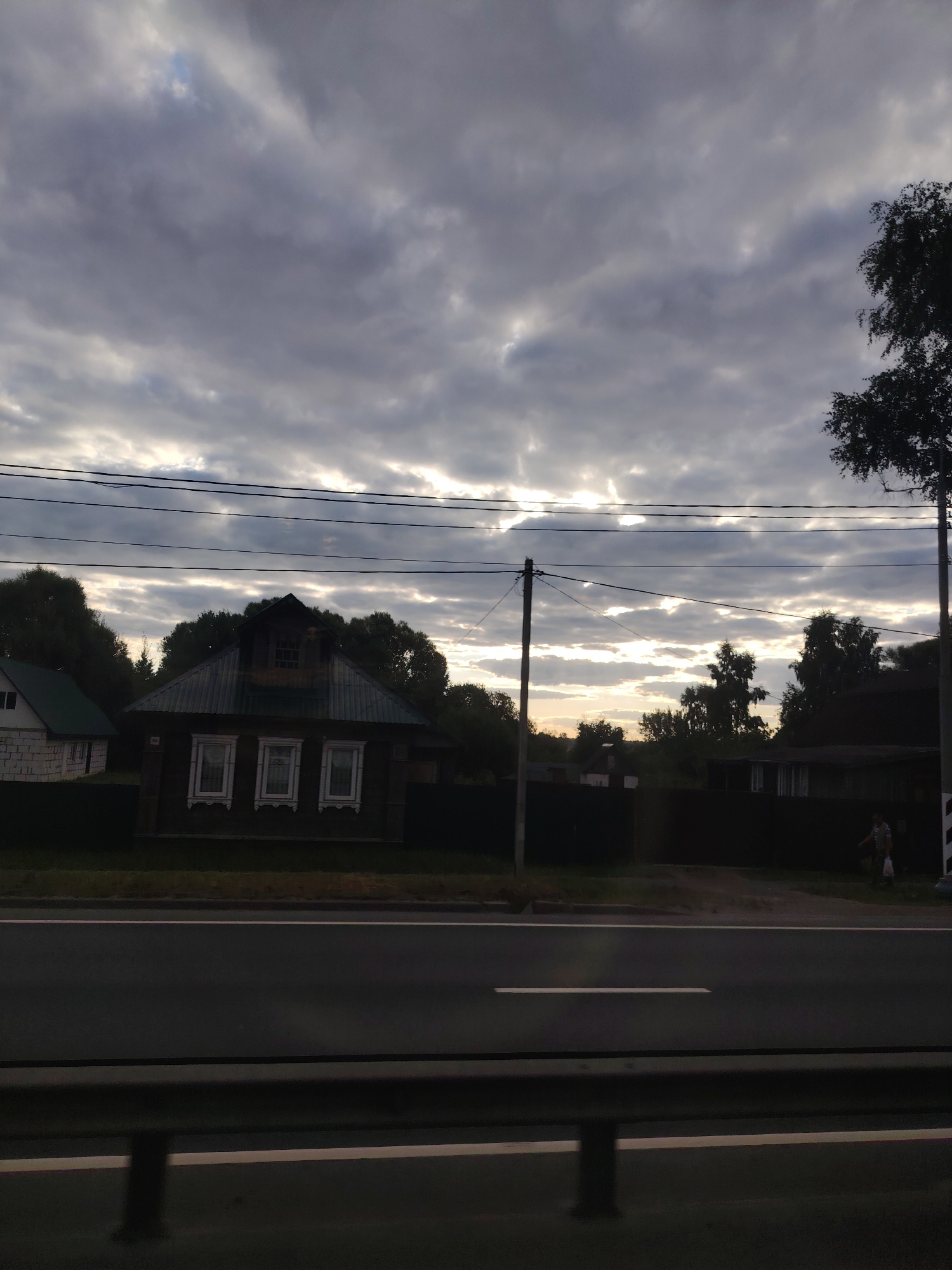The image shows a rural landscape, possibly in the city of Spas-Zaulok, Russia, under a cloudy sky. In the foreground, a dark asphalt road runs horizontally across the frame, with a guardrail visible in the bottom section. Beyond the road, a dark fence encloses several properties. To the left, a dark wooden house with three white-trimmed windows and a dormer window is prominently featured. To the right of this house, a lighter colored building with a green roof is partially visible. Further to the right, silhouetted trees and other structures are present. A single person is seen walking on the right side, carrying a bag. Overhead, power lines are strung across the sky. The sky is dramatic, with patches of sunlight breaking through dense, dark clouds.