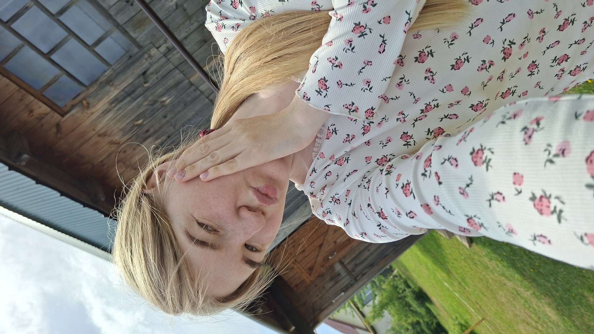 A person with long blonde hair and wearing a white shirt with pink flower patterns is partially obscuring their face with their right hand. The location appears to be in Большой Букор, Russia, as suggested by the query. The background includes parts of a wooden structure, potentially a building or fence, and a glimpse of green grass and sky. The image is oriented in a rotated perspective.