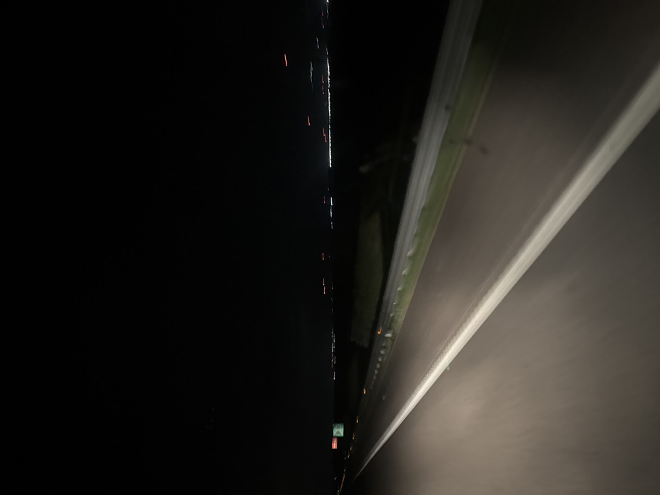 The image depicts a nighttime view from a moving vehicle on a multi-lane highway, likely an autostrada, located in an outlying area of Foggia, Italy. The sky above is uniformly dark. The asphalt road surface, illuminated by the perspective vehicle's headlights, features a prominent solid white lane marking along its immediate left edge, and a less distinct white line on the right delineating the shoulder. A metal guardrail with reflective markers runs along the right side of the road, separating it from a dark, grassy strip of land. A similar guardrail is partially visible further left, suggesting a median or opposing traffic lanes. In the far left distance, a green highway sign with unreadable white text is present. The distant horizon is marked by a blurred horizontal array of white and red artificial lights, indicative of a city or town, consistent with the urban periphery of Foggia, Italy. No individuals are visible. The scene implies vehicular travel at speed during darkness.