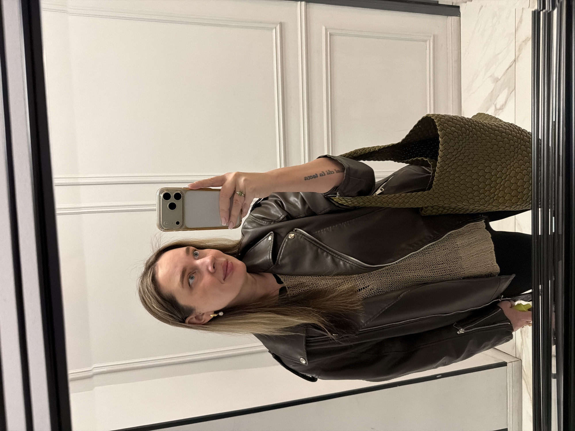 A woman takes a mirror selfie indoors, in a setting reportedly located in Moscow, Russia. She is reflected facing slightly to her left, looking forward with a neutral expression and a faint smile. She has light-colored hair, which falls past her shoulders, and is wearing a dark brown or olive-green leather-look motorcycle jacket over a light beige or tan knitted top with a visible mesh texture. A pearl earring is visible in her right ear.
She holds a gold-colored iPhone with three camera lenses in her left hand, which is raised to capture the reflection. On her left ring finger, she wears a ring with a light green stone. Her right forearm features a tattoo with text that appears reversed in the mirror, reading "rual al ihd alu". An olive-green, textured tote bag, possibly made of woven or quilted neoprene, hangs from her right shoulder.
The background shows a well-lit room with white walls featuring classic decorative molding and paneling. To the right of the reflection, a section of the wall displays white and grey marble patterning. The floor is light-colored, possibly tiled. The selfie is taken in a mirror with a prominent black frame. The left side of the frame has vertical black and white stripes, while the right side is plain black.