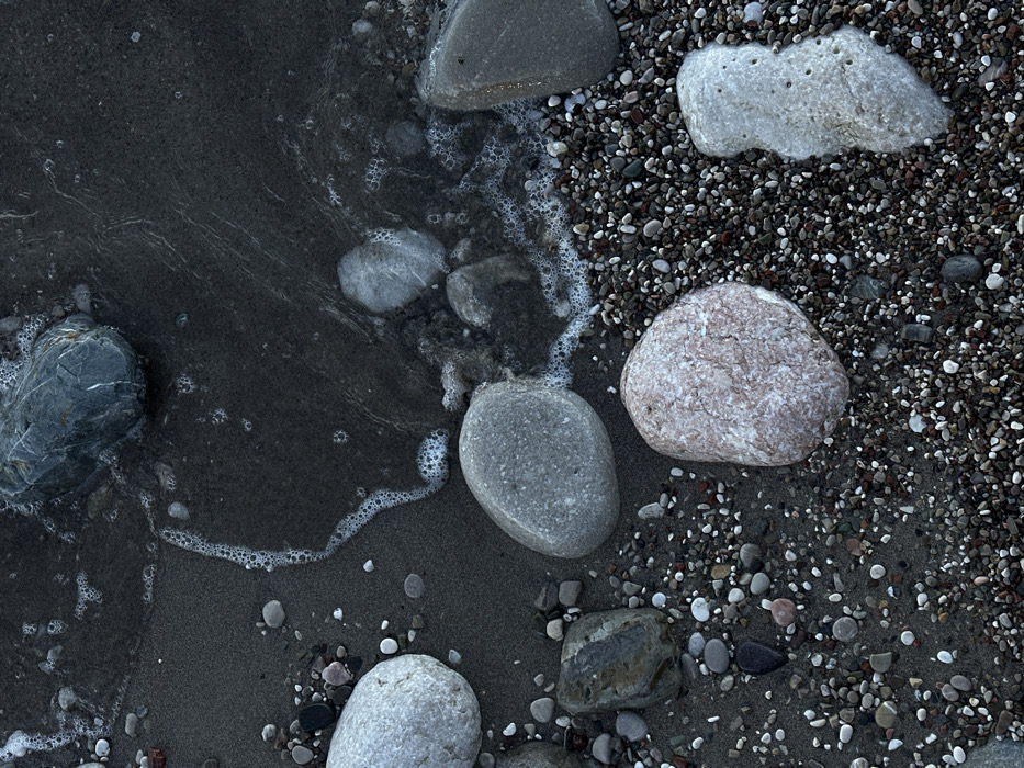 This image presents an overhead, close-up view of a pebbly beach at the water's edge in Bar, Montenegro. The scene is dominated by dark, wet sand intermixed with a dense collection of small, multi-colored pebbles in shades of black, grey, white, and reddish-brown. Numerous larger, rounded stones are scattered across the frame, including a prominent light grey stone on the upper left, a darker grey stone partially submerged in water at the upper right, a medium grey stone near the center, a reddish-pinkish stone in the lower-middle, and an irregularly shaped, porous off-white stone on the lower right. Dark water occupies the upper portion of the image, actively washing over the sand and pebbles, evidenced by white foam lines and clusters of small bubbles marking the water's receding or advancing edge, particularly a distinct, curving trail extending from the upper-left towards the center-right. No individuals or man-made objects are visible. The lighting appears dim, suggesting low-light conditions.