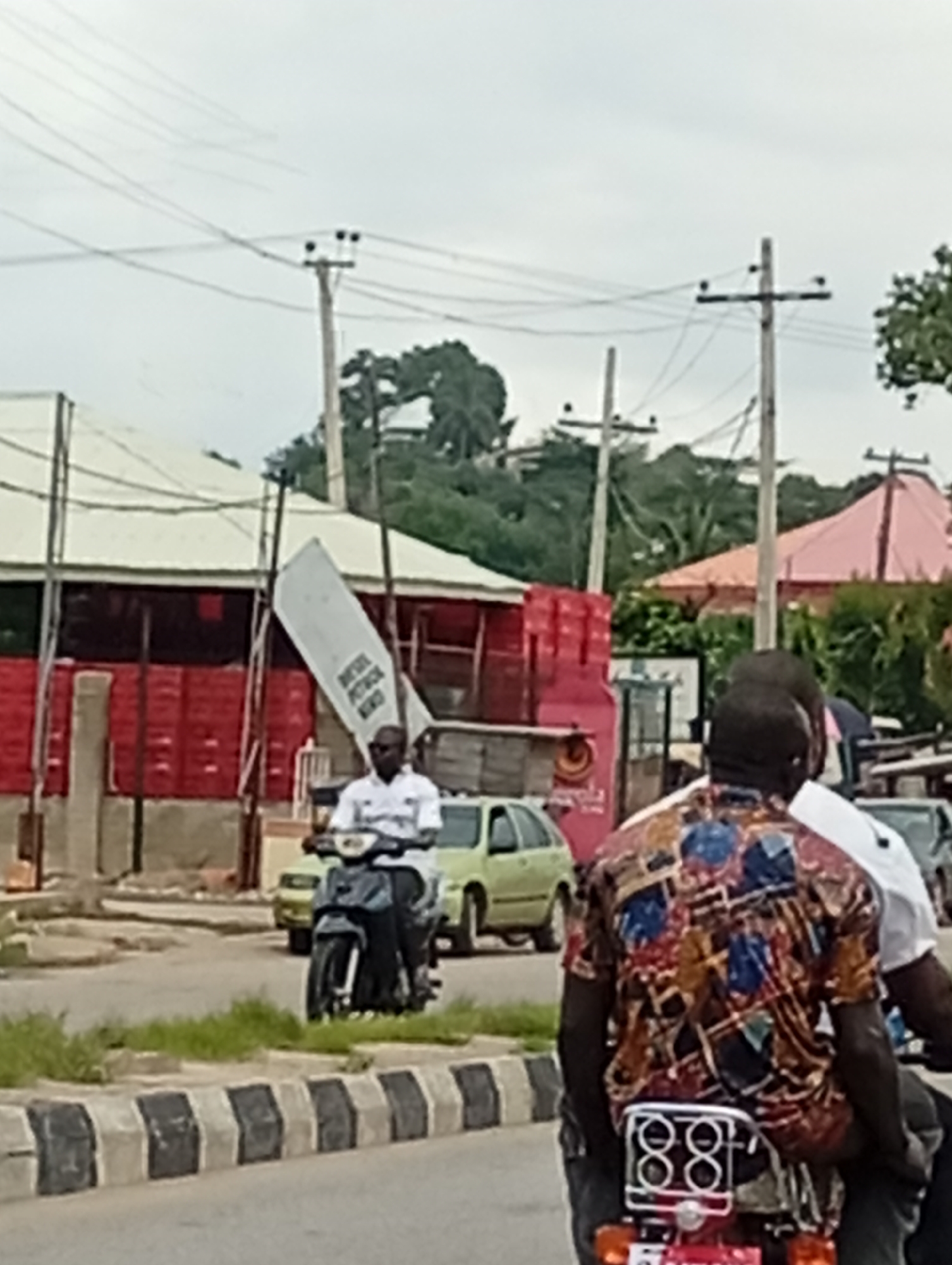 In an outdoor urban environment, the image captures a cloudy day with a street scene. A man is riding a motorcycle, wearing a white shirt and sunglasses. Another person, wearing a colorful patterned shirt, is on the back of the motorcycle. In the background, there are buildings with red and white facades, trees, and power lines. A signpost leans precariously against one of the buildings. A light green car is parked on the side of the road, and another vehicle is partially visible behind it. The foreground features a striped concrete barrier along the road. The overall impression is a busy street with typical urban elements and activities.