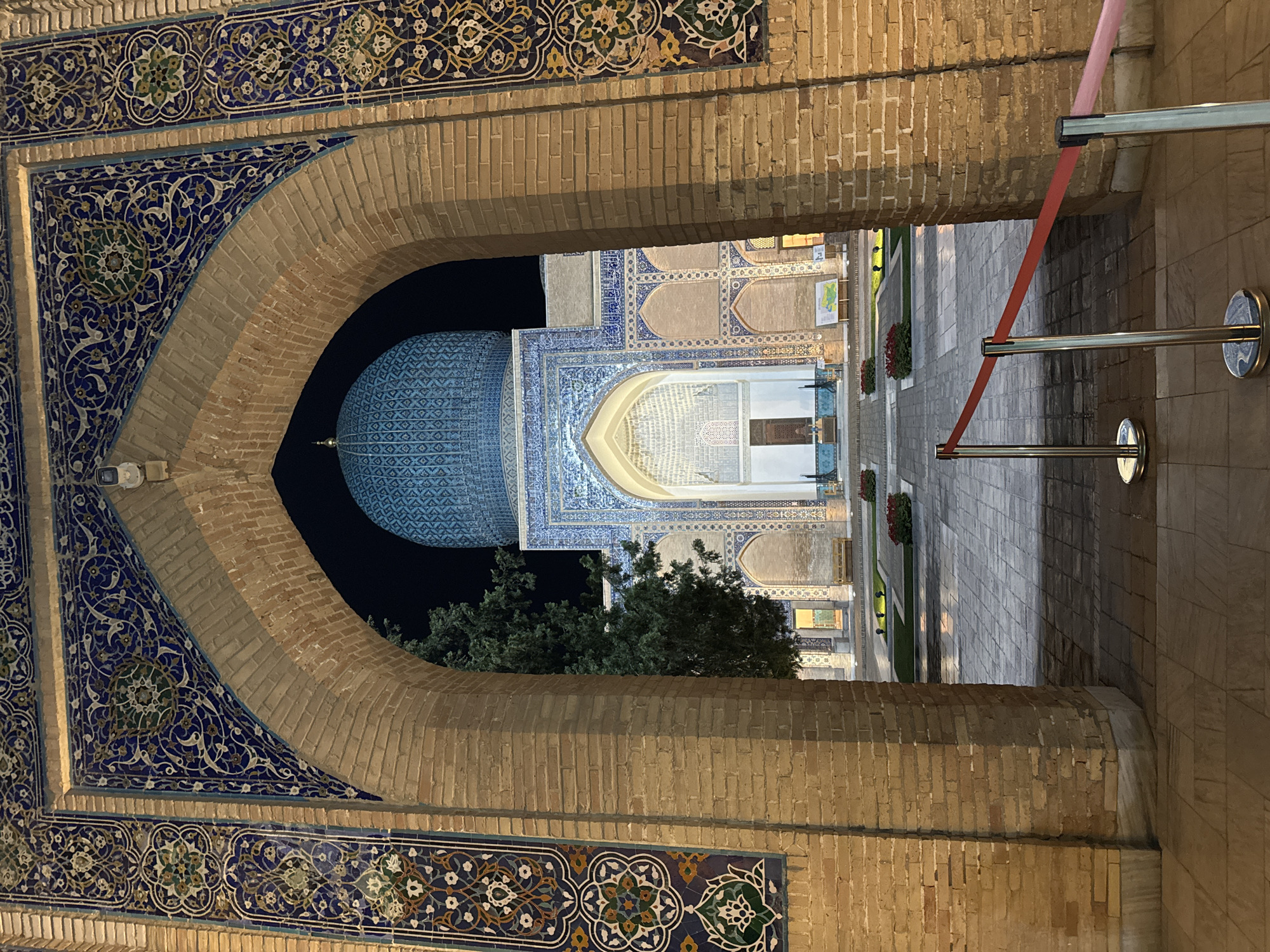 The image depicts a view of the Gur-e-Amir Mausoleum in Samarkand City, Uzbekistan, as seen through an arched doorway. The archway is made of bricks and decorated with intricate blue and gold tilework. Through the arch, the mausoleum is visible, featuring its iconic blue dome, tiled facades, and a gated entrance. A paved courtyard lies in front of the mausoleum, lined with greenery and flowerbeds. A red rope barrier with metal stanchions is positioned on the right side of the archway, limiting access. The sky is black behind the dome, suggesting nighttime or a shadowed area. Some trees are also visible.