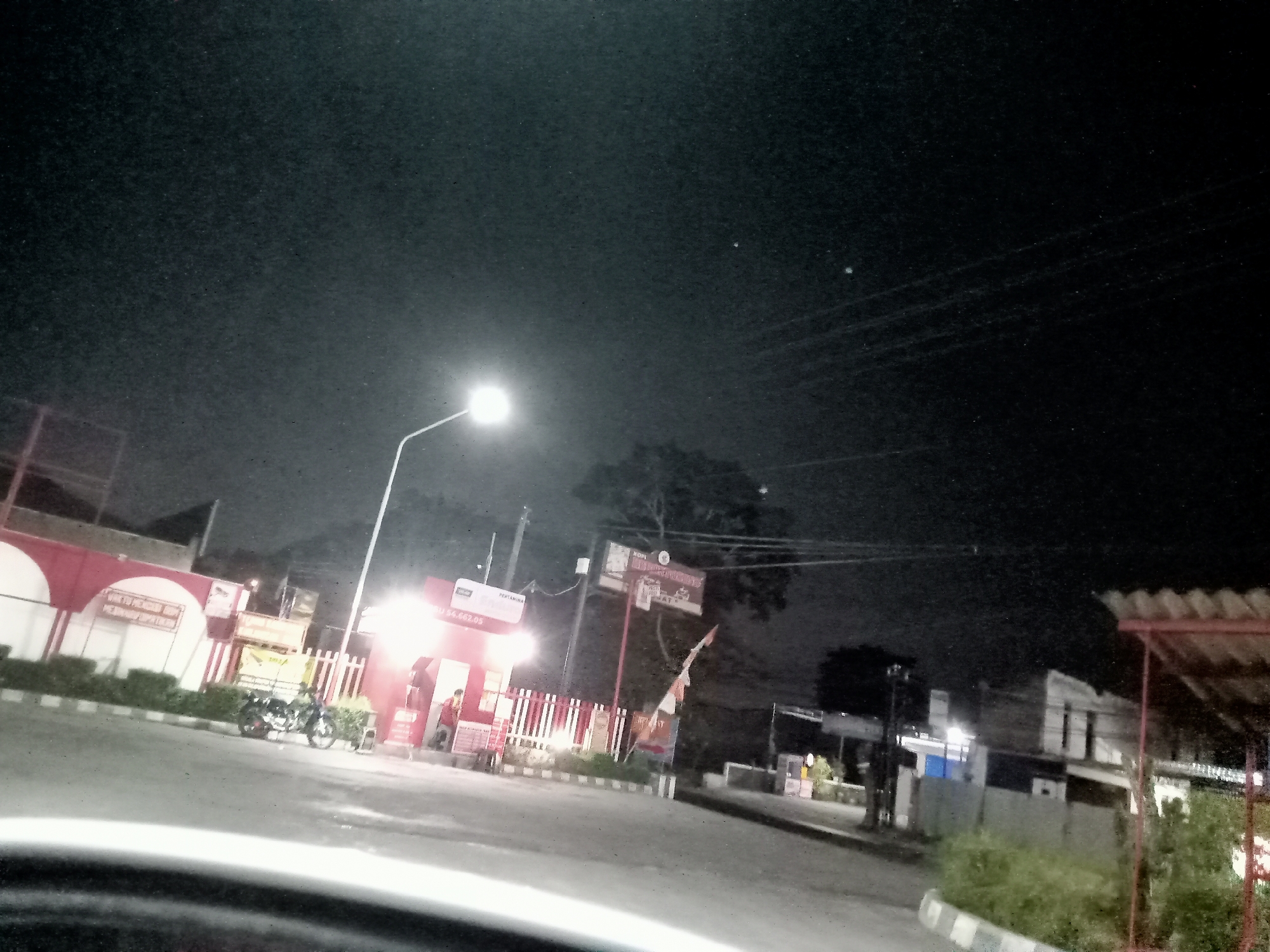 The image depicts a nighttime scene at a roadside establishment, likely a gas station or convenience store, in Mangunsari, Indonesia. A bright streetlamp illuminates the paved road and the front of the building, which features red and white signage. A motorcycle is parked in front of the establishment. A person, dressed in red, is visible inside the building. The sky above is dark, with a few faint lights visible in the distance. The overall impression is one of a typical urban setting at night, with artificial lighting casting shadows and highlighting the immediate surroundings.
