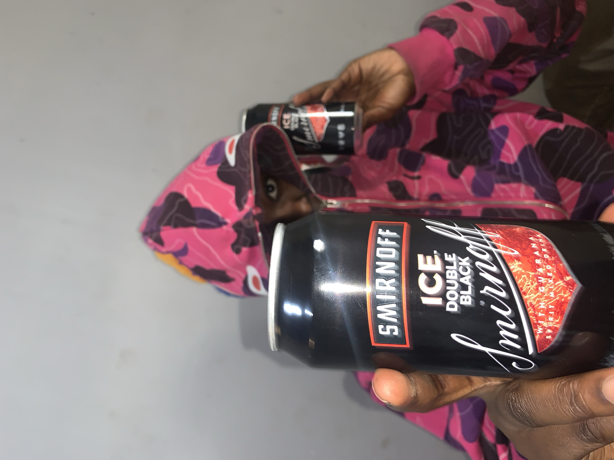 A person wearing a pink and black camouflage hooded sweatshirt is holding two cans of Smirnoff Ice Double Black alcoholic beverages. The scene's location and context within Rafin Par, Nigeria, are not provided. The background consists of a grey wall.