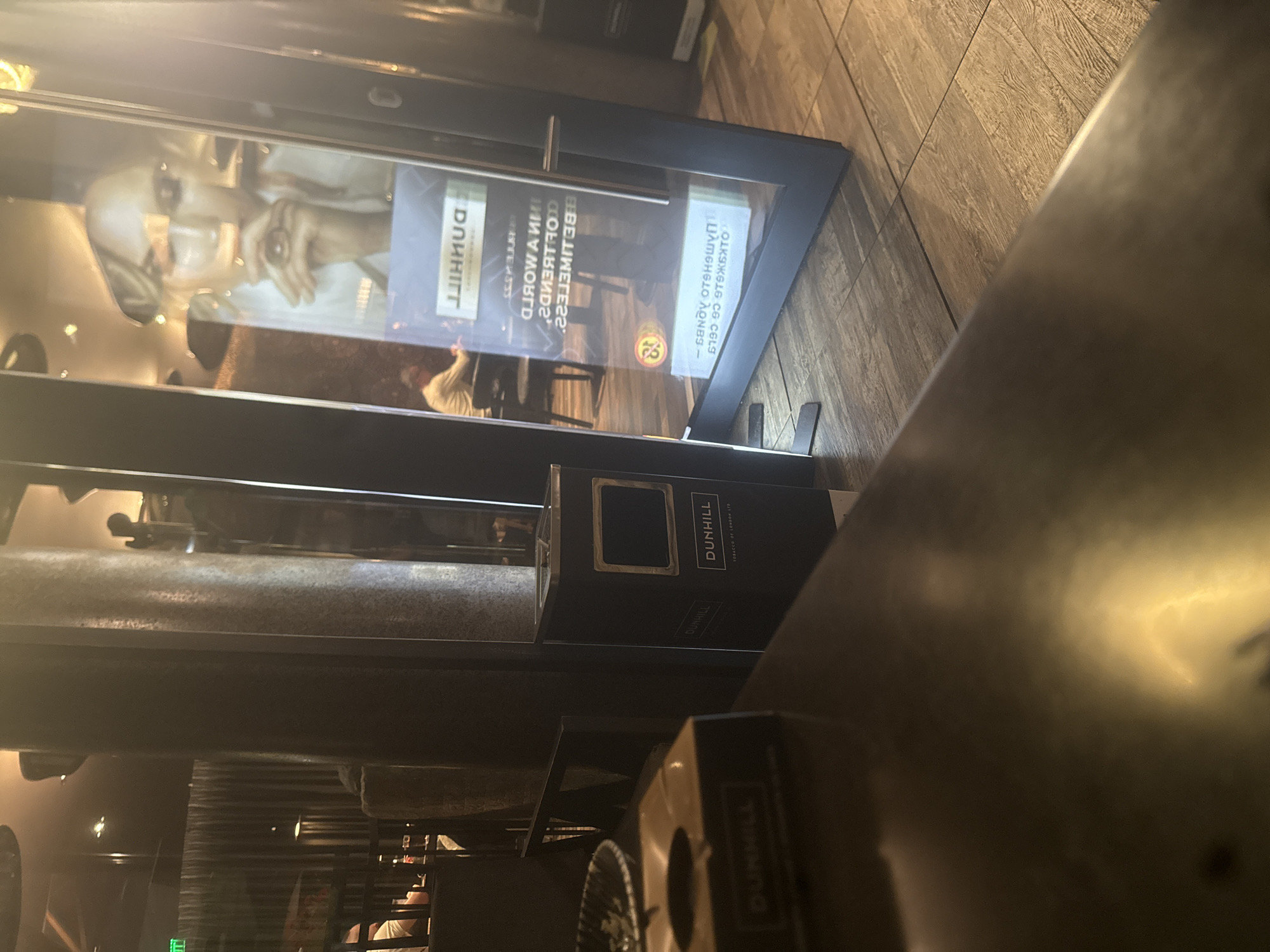 This image depicts a dimly lit, high-end indoor establishment in Sofia, Bulgaria, likely a lounge or smoking area. In the immediate foreground, a dark, reflective tabletop holds a black, square Dunhill-branded ashtray. To the right, a black Dunhill-branded bin/urn with a silver top, displaying "DUNHILL TOBACCO OF LONDON LTD," rests on a light brown, wood-grain tiled floor.
In the mid-ground, a prominent vertical display panel reflects the interior. The reflection prominently features an inverted image of a blonde woman, with "DUNHILL" branding above inverted Cyrillic text, including "ПУШЕНЕТО УБИВА" (Smoking Kills). Reflected within this panel, a person wearing a light-colored top is seated at a table further back in the room. Another person is partially visible in the far left background, also seated.
The background shows dark, textured pillars, subtle lighting fixtures on a dark ceiling, and more dark furniture, suggesting a sophisticated atmosphere. A green "EXIT" sign is visible in the upper left background.