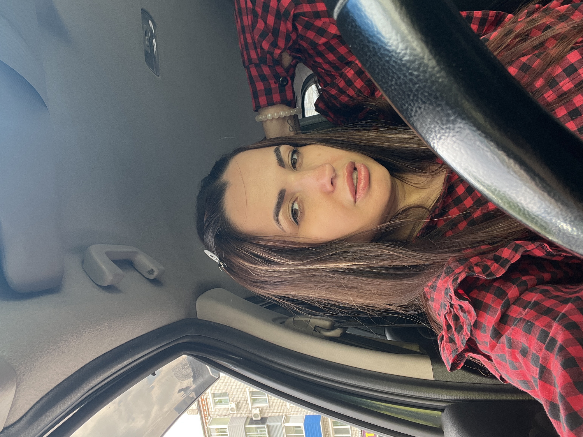 A selfie-style photograph taken inside a car, oriented sideways. The subject, a woman with long brown hair, occupies the center of the frame. She is wearing a red and black plaid shirt. A steering wheel is visible in the foreground. Through the car window, a building with multiple windows and air conditioning units is visible, suggesting an urban environment. The location is Unknown, Unknown.