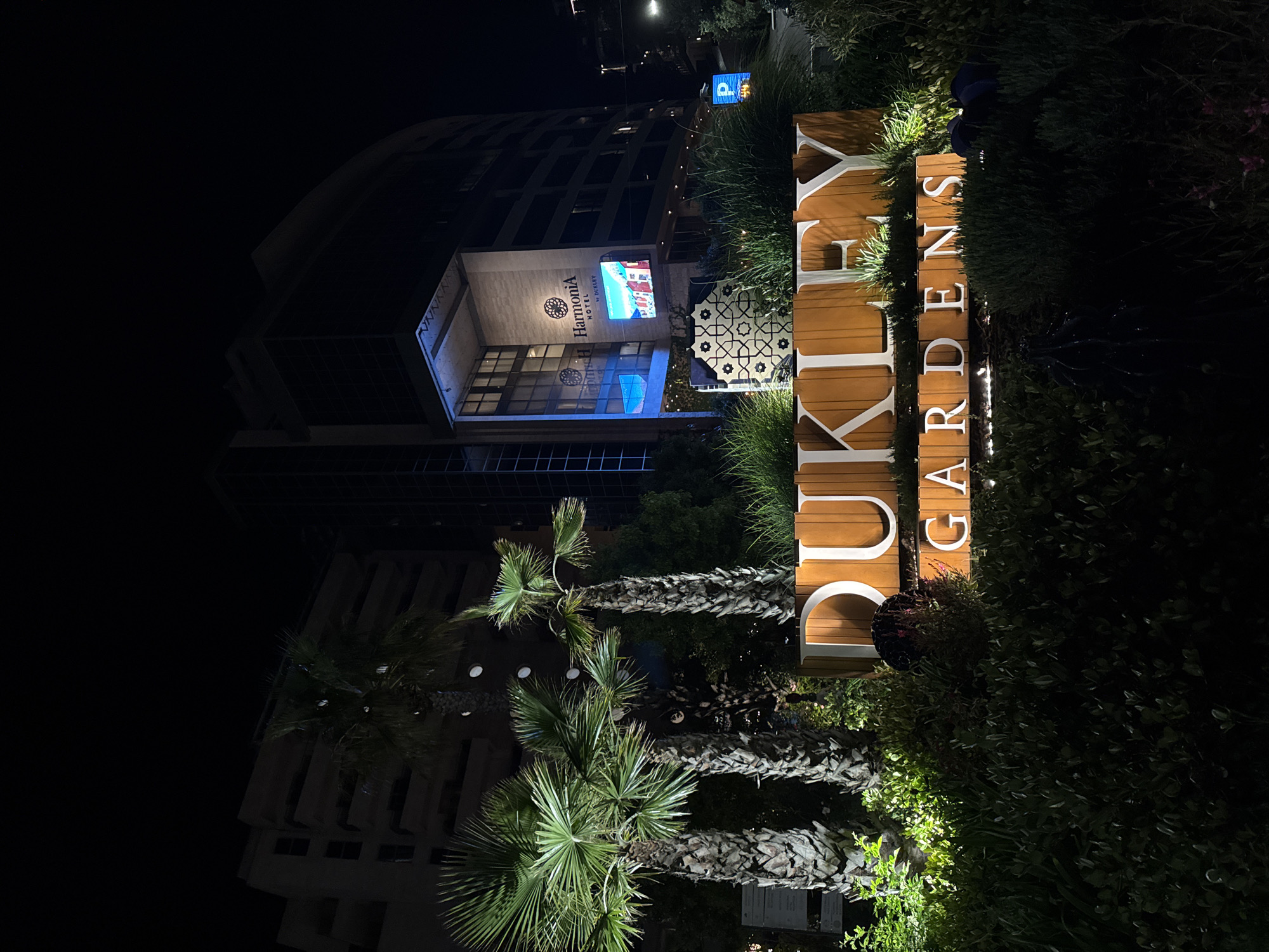 At night in Budva, Montenegro, a vertical sign reading "DUKLEY GARDENS" is prominently displayed. The sign is constructed of wood and illuminated from within, making the white letters stand out. It is positioned in front of dense greenery and some palm trees, suggesting a garden or landscaped area. Behind the sign, the Harmonia Hotel is visible, illuminated with exterior lighting and featuring a large digital screen displaying an image. The hotel appears modern and multi-storied. The sky is dark, and streetlights are visible in the background.