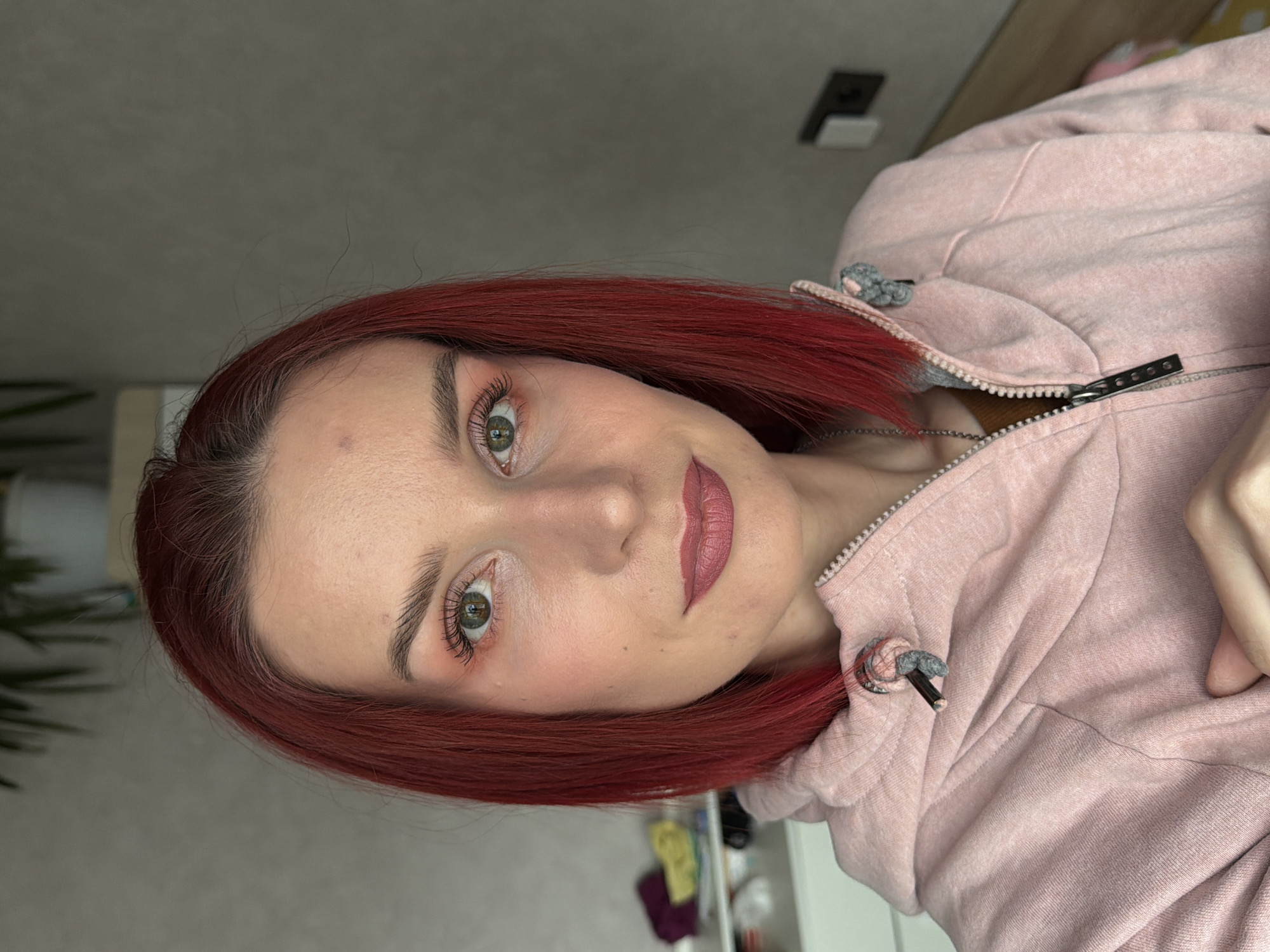 The image is a selfie of a fair-skinned woman with dyed red hair. She has green eyes and is wearing makeup, including lipstick, blush, and eye makeup. She is wearing a light pink hoodie. The background is a gray wall, a light-colored wooden surface, and a glimpse of a white shelf unit with various items on it. There is also a light switch visible on the wall. There is no information in the image to establish that the photo was taken in Chaikovsky, Russia.