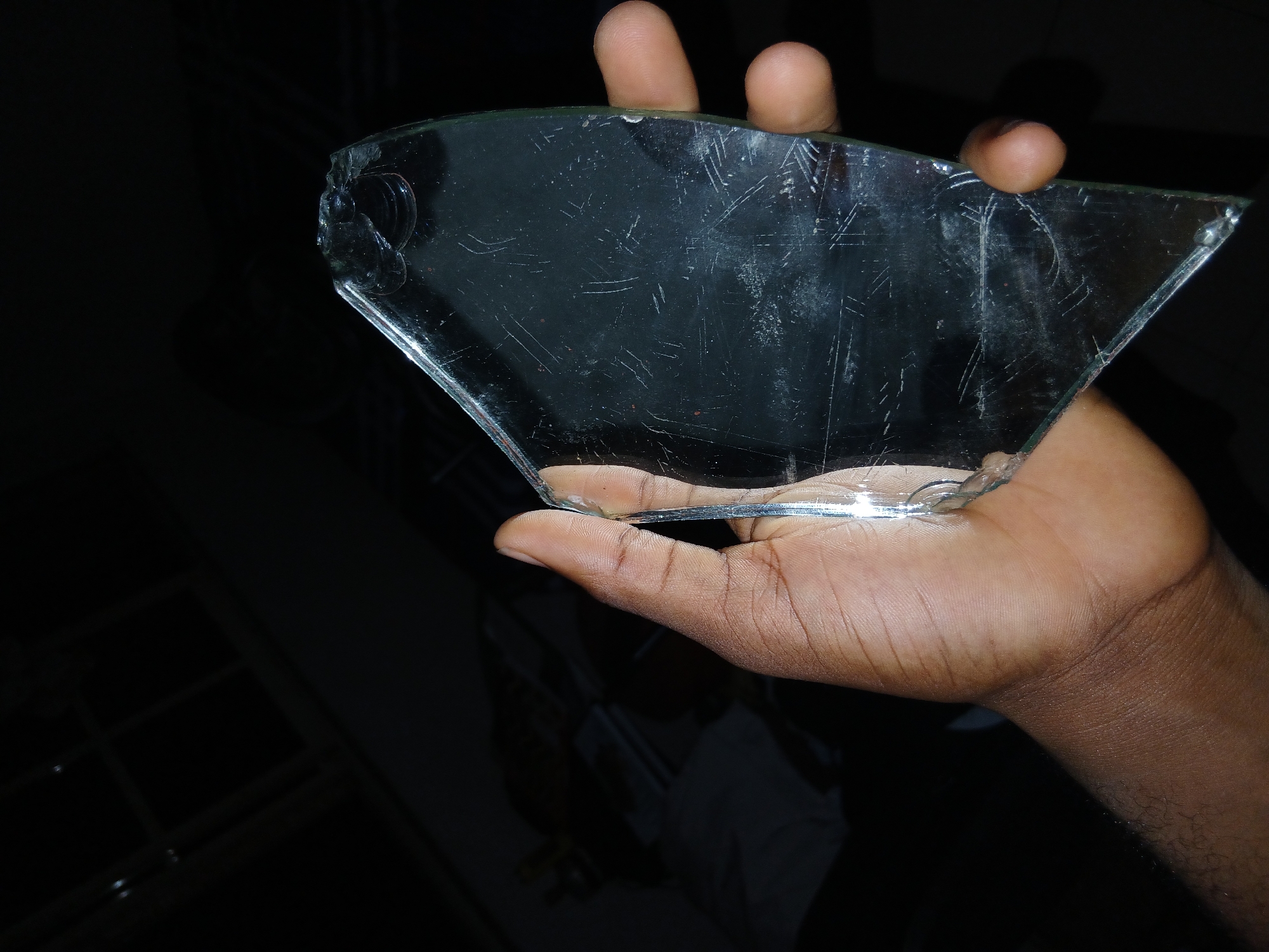 An adult's right hand with medium to dark brown skin tone is prominently featured, holding a large, irregularly shaped piece of broken transparent glass against a predominantly dark, indistinct background. The hand is positioned centrally, with fingers curved around the glass, supporting it from below and the sides.
The piece of glass is roughly trapezoidal or fan-shaped, with jagged, sharp edges typical of broken glass. It is heavily scratched and appears somewhat smudged or dirty. A small, circular indentation or hole is visible near one of the broken edges on the left side, and some translucent, glue-like residue is present along another edge. The glass reflects light, creating bright highlights on its surface and edges, emphasizing its sharp features.
The background is almost entirely black, suggesting a dimly lit indoor environment or nighttime. Faint, dark outlines of what might be window panes or a grid-like structure are barely discernible in the lower left, but provide no specific location cues. No explicit activity is depicted beyond the act of holding the glass, and no emotions are discernible from the hand. There is no visible text in the image.
Given the location context of Lagos, Nigeria, the image itself does not provide specific visual information to confirm this geographical location.