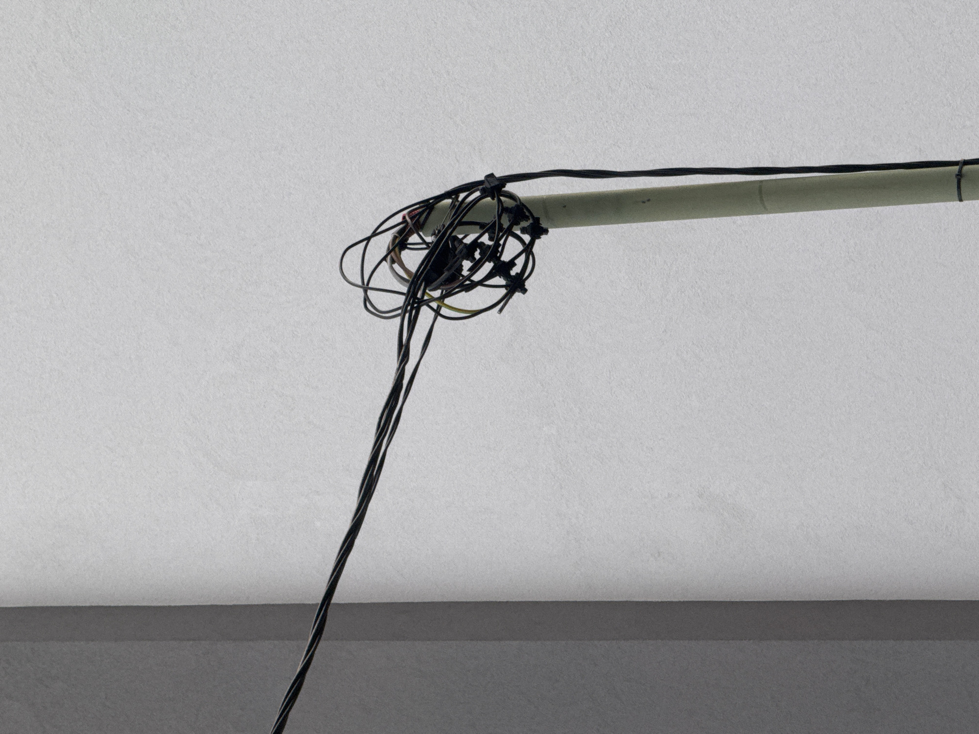 The image displays a utility pole in Sofia, Bulgaria, against a textured gray wall. The pole is topped with a tangled mass of black electrical wires, some of which extend out of the frame. A single black wire hangs down from the cluster of wires, secured near the base of the pole with a black zip tie. The lighting suggests an overcast day.