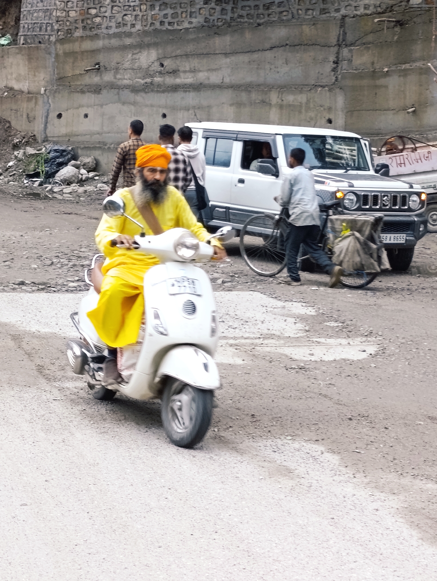 In Manali, India, a Sikh man wearing a yellow turban and traditional yellow attire rides a white scooter. He has a long, dark beard and is holding the scooter's handlebars. Behind him, a white SUV is parked, with its front grill visible. A person in a light-colored shirt and dark pants is near the SUV, pushing a bicycle with a large cloth-covered carrier. In the background, other individuals are visible, one wearing a plaid shirt and another a hooded sweatshirt. A concrete wall with stone detailing forms the backdrop, and piles of debris and rocks are present near the wall. The ground is a dirt and gravel road.