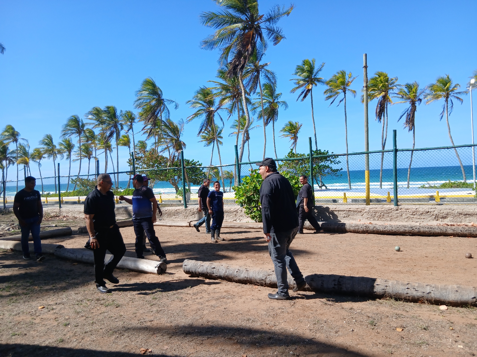 The image captures an outdoor scene on a bright, sunny day, likely in a tropical or coastal region, specifically Playa El Agua, Venezuela, as indicated by the context. Several adult men are gathered on a dirt court, engaged in what appears to be a casual game similar to bocce or petanque.
In the foreground and midground, approximately seven to eight men are visible. They are casually dressed in t-shirts, polo shirts, jeans, and closed-toe shoes. One man on the left wears a black t-shirt with "Nautica" printed on it. Another man, central to the image and facing away from the camera, wears a black polo shirt and dark pants. To his right, a man in a blue t-shirt with "Champion" visible is smiling towards the background. Most men are standing or walking within or around the playing area, suggesting active participation or observation of the game.
The playing surface is unpaved, consisting of packed dirt or sand. Long, weathered logs, likely palm tree trunks, define the boundaries of the playing area. Two small, round balls – one green and one brownish-grey – rest on the dirt, indicating the game in progress.
The background is dominated by a clear, vibrant blue sky and numerous tall, slender palm trees with green fronds, some swaying slightly, hinting at a breeze. Beyond the playing court and palm trees, a chain-link fence separates the area from a concrete structure with yellow and white markings. The bright blue ocean with white waves crashing on the shore is clearly visible behind the fence and concrete barrier, further emphasizing the beachside setting. A utility pole or light post is also visible among the palm trees.
The scene conveys a relaxed, recreational atmosphere under brilliant sunshine, likely in the mid-morning or early afternoon.