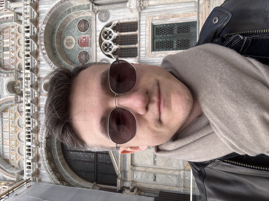 A male individual is depicted in a close-up, chest-up self-portrait, positioned centrally. He has short, dark hair, wears oval-framed sunglasses with dark lenses, and exhibits a faint smile. His attire consists of a dark leather jacket and a light beige scarf wrapped around his neck. The background is the ornate facade of St. Mark's Basilica in Piazza San Marco, Venice, Italy. Visible architectural details include multiple marble arches, decorative columns, windows with dark grilles, and mosaic elements in various colors of stone and glass, consistent with the Byzantine-Gothic style of the basilica.