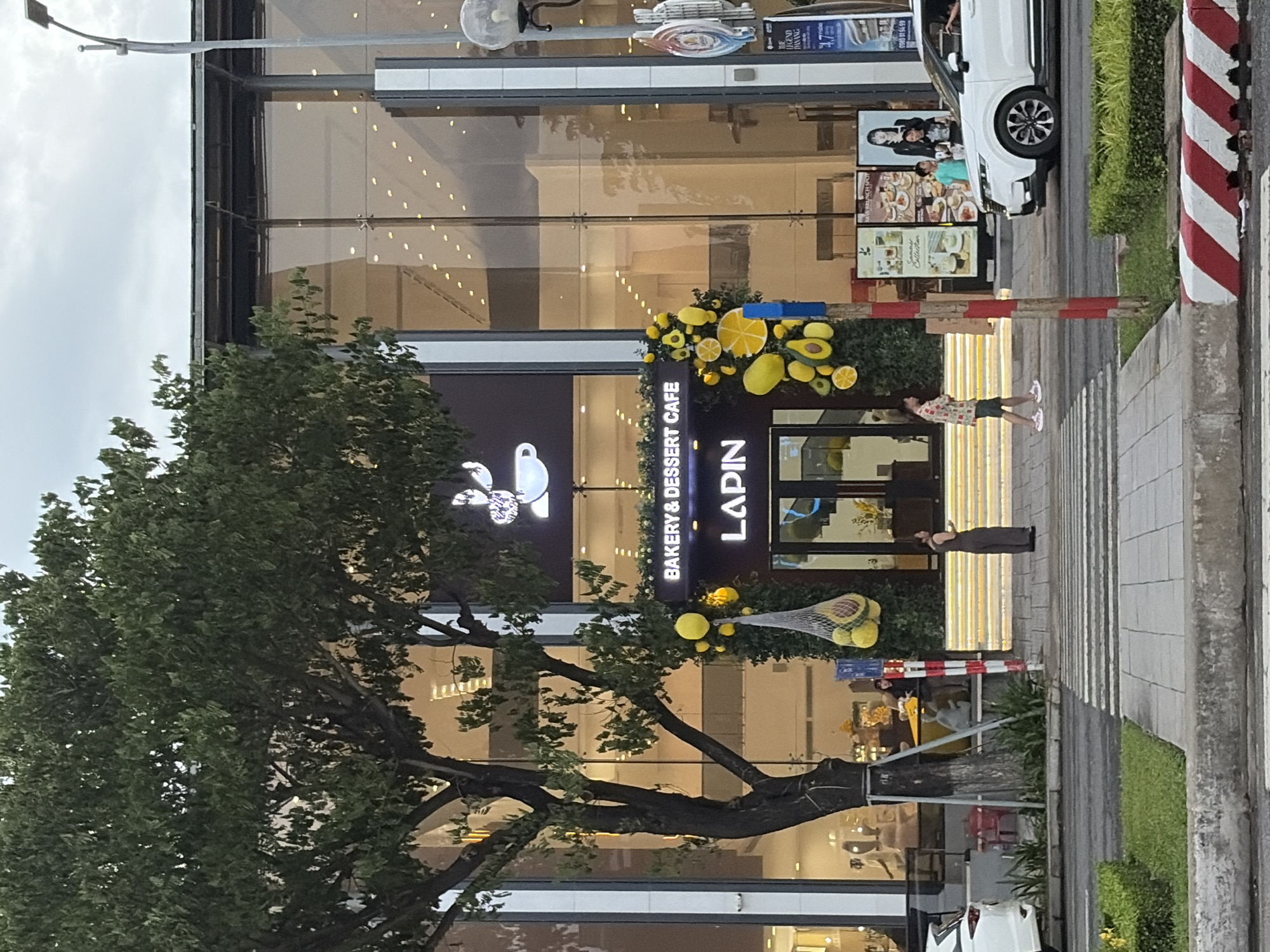 The image depicts the exterior of a bakery and dessert cafe named "LAPIN" in Đà Nẵng, Vietnam, on an overcast day. The cafe has a modern facade with large glass windows reflecting the cloudy sky. A prominent tree with dense green foliage stands to the left of the entrance. The entrance itself is decorated with yellow balloons and artificial fruits, including lemons, avocados, and slices of citrus.
Several individuals are visible. A woman in a long, dark dress stands on the illuminated steps leading to the cafe entrance. To her right, a young man wearing a patterned shirt and shorts walks across the zebra crossing. Further to the right, a white SUV is parked near the curb, and two people are seen inside it. In the background, posters advertising menu items and a woman are displayed on the building's exterior. The street in front of the cafe has a zebra crossing, and a concrete curb with red and white markings runs along the road. Green grass and hedges line the sidewalk. The overall scene suggests a street-level view of a commercial establishment in an urban environment.