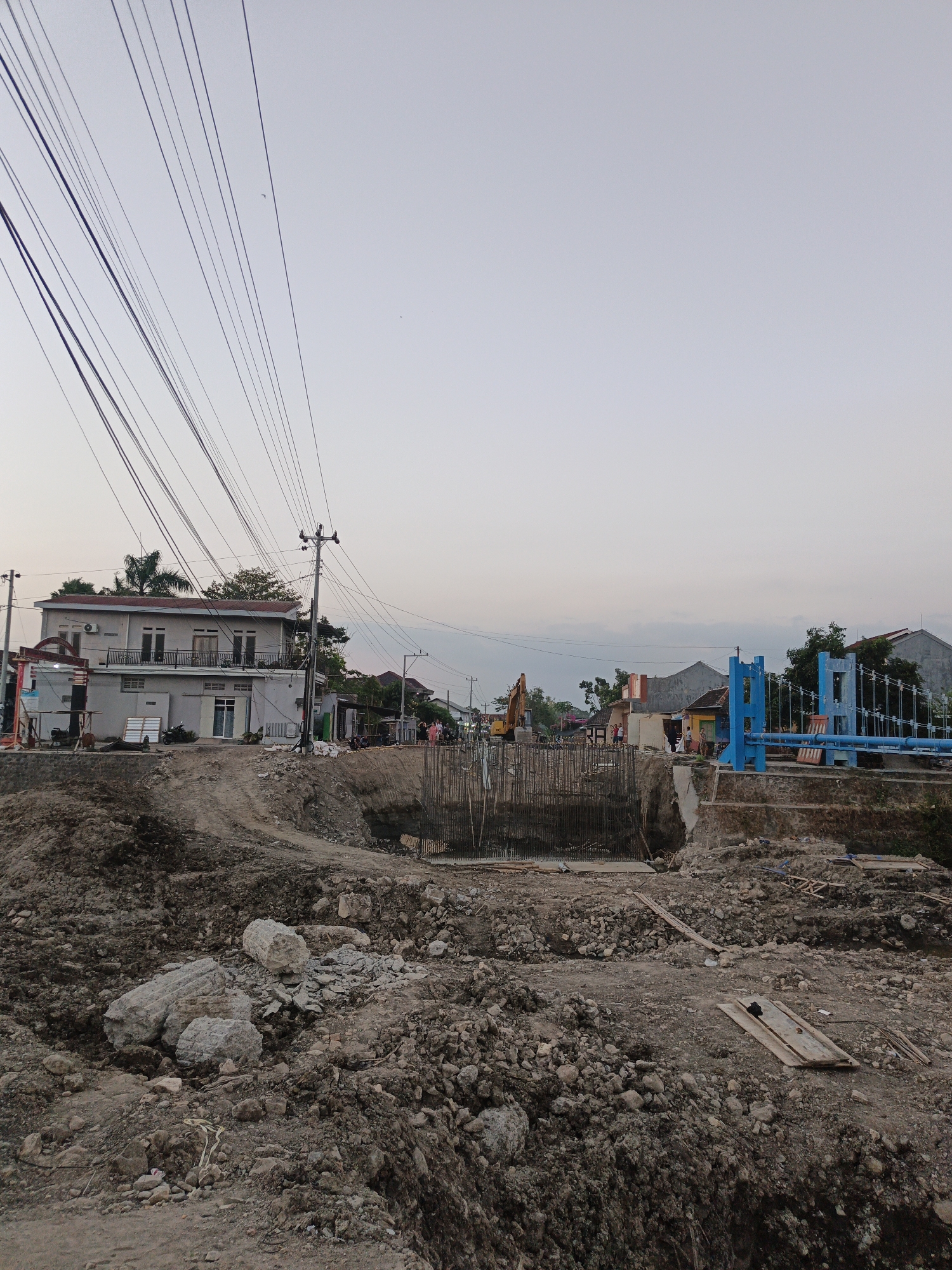 The image depicts an active construction site in Planggu, Indonesia, focusing on the development of a bridge or overpass. The foreground is dominated by a large area of disturbed earth, characterized by mounds of soil, rocks, and debris, indicative of excavation work. Several large stones, one appearing to be a broken concrete block, are scattered across the uneven ground.
In the mid-ground, the construction of a structure with visible rebar and concrete foundations is underway in a dug-out area, likely for the bridge's support. To the right, a bright blue suspension bridge stands completed or near completion, with its distinctive support towers and cables. An excavator is visible further back, suggesting ongoing heavy machinery operations.
Several buildings are situated along the periphery of the construction site. A prominent two-story building with balconies stands on the left, with numerous power lines converging overhead. Other residential structures with varying rooflines are visible in the background, interspersed with trees. A few individuals are present, appearing to be workers or onlookers, observing the construction activities. The sky is overcast with a pale, diffused light, suggesting either early morning or late afternoon. The overall scene captures a transition from raw earth to developing infrastructure within a semi-urban or rural environment.