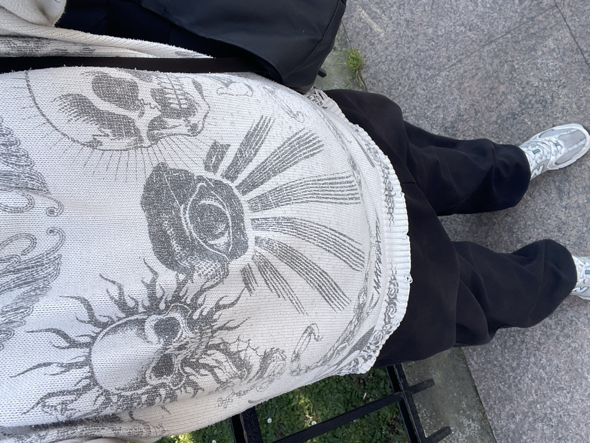 The image displays a downward-angled view of an individual standing outdoors in Budva, Montenegro. The person is wearing a light-colored, likely white or off-white, long-sleeved knitted sweater adorned with dark grey intricate designs. Central to the sweater's pattern is an eye emitting radiating lines, flanked by two distinct skull motifs. The skull on the viewer's left features prominent flame-like or sun-like rays, while the skull on the right is depicted without such embellishments. Additional abstract patterns and partial illegible text are visible near the ribbed bottom hem of the sweater. The individual wears dark, possibly black, baggy trousers and light-colored athletic shoes with grey and light blue accents. A black bag hangs vertically on the person's right side. The ground beneath the person is a light grey, speckled paved surface, consistent with a sidewalk or pedestrian path. To the left, a segment of green grass with small white flowers is visible, separated from the paved area by a black metal fence or railing. The scene is illuminated by natural daylight.