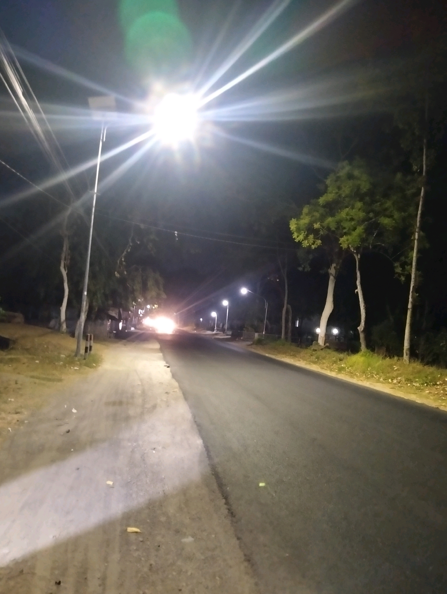 The image depicts a nighttime street scene in Wonoketro, Indonesia, illuminated by a bright street light in the upper center of the frame. The street light casts a strong glare, creating visible light rays that extend outwards. A paved road occupies the right side of the image, stretching into the distance. To the left of the road, there is a dirt or gravel path. Several trees line the sides of the road, their leaves appearing green in the artificial light. A few smaller street lights are visible further down the road. In the background, there is a vehicle with its headlights on, creating a bright white light. The overall environment appears to be a rural or suburban road at night.