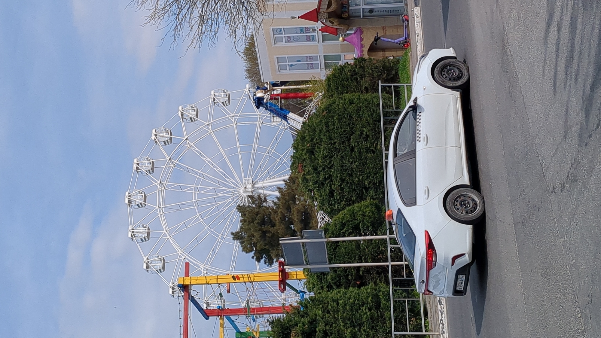 This daytime image captures a vibrant scene at an amusement park adjacent to a road, likely in Gelendzhik, Russia. A large, white Ferris wheel dominates the left side of the frame, its many gondolas silhouetted against a bright blue sky with scattered clouds. Below the Ferris wheel, parts of other colorful rides are visible, including a red and blue structure and a yellow and red base, suggesting a lively recreational area.
In the mid-ground, dense green bushes and trees provide natural landscaping, partially obscuring the base of the rides. A light-colored building with large windows stands behind the greenery, possibly part of the park or a nearby establishment. Cyrillic text is visible on the building, including "МЕД ЦЕНТР" (Med Center) and "ХОЛТЕР" (Holter), indicating a medical facility. A purple scooter is parked near the building.
On the right, a white sedan, identifiable as a taxi by a checkered strip on its side and an orange light on its roof, is parked or driving on an asphalt road. The car's rear license plate is partially visible, displaying "P194AE 793". The region code "793" corresponds to Krasnodar Krai, confirming the provided location context of Gelendzhik, Russia. Bare tree branches are visible in the upper left corner against the sky. The scene is static, with no explicit activity, presenting a snapshot of the park and its surroundings under clear weather conditions.