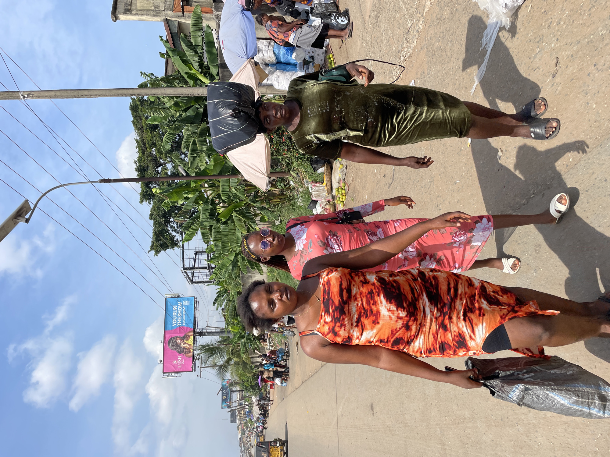 The image captures three women standing on a street in Onitsha, Nigeria. The woman in the foreground wears a sleeveless orange and white tie-dye dress and carries a dark, shiny bag in her right hand. The woman behind her is dressed in a pink floral print dress and has sunglasses perched on her head. The third woman, positioned furthest back, is wearing a green velvet dress and has a large black bag on her head. They appear to be walking or posing on a paved street.
The background suggests a bustling outdoor market scene with numerous people and various goods, including bananas and what appear to be other produce items. Several umbrellas are visible, indicating a sunny day or protection from the sun. In the distance, buildings and a large billboard are present. The billboard displays text that includes "YOUR RUN THE SHOW" and "BE CONSISTENT," along with images of people. The lighting suggests it is daytime, likely mid-morning or afternoon, given the clear sky and shadows. The overall atmosphere seems active and typical of a busy Nigerian street market.