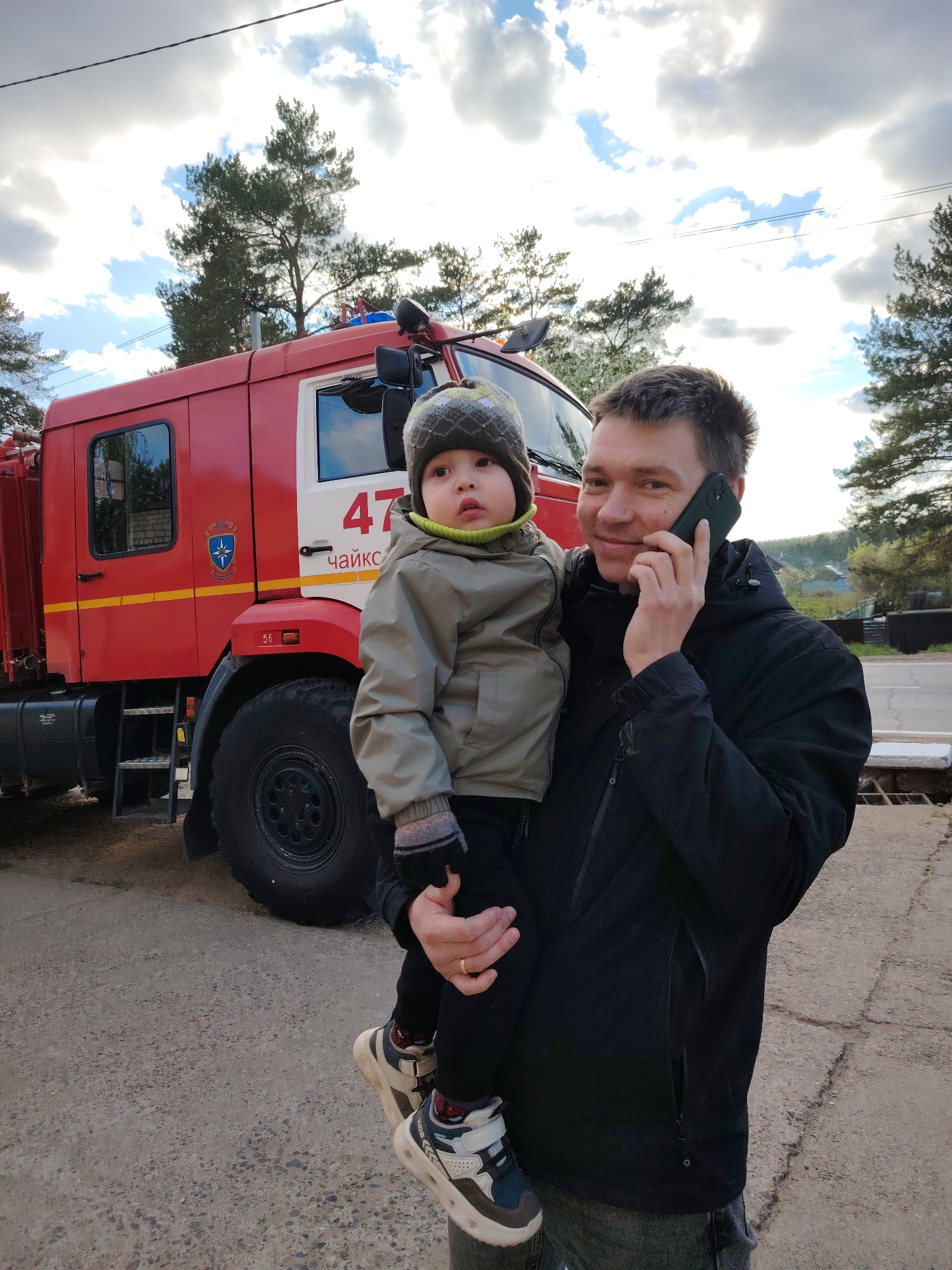 A man is holding a young child in front of a red fire truck. The man is wearing a black jacket and jeans, and he is talking on a cell phone. The child is wearing a green jacket, a hat, and sneakers. The fire truck has the number "47" and the word "Чайковский" (Chaikovsky) on the side. The background includes trees, a road, and a partly cloudy sky. The scene appears to be in or near the city of Chaikovsky, Russia.