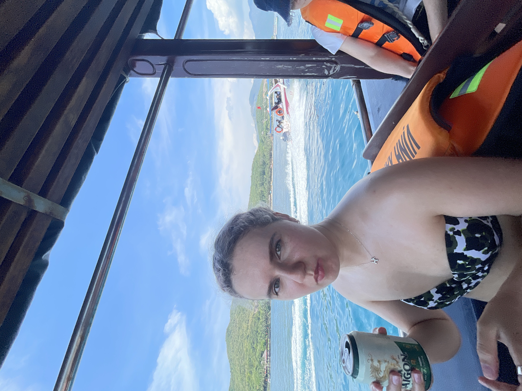 A woman holds a can of Saigon beer on a boat, wearing a swimsuit, necklace, and a lifejacket nearby, with a view of the ocean, a smaller boat in the distance, and green, mountainous land near Nam Nha Trang Ward, Vietnam. The shot is taken from under the boat's canopy, showing the sky.