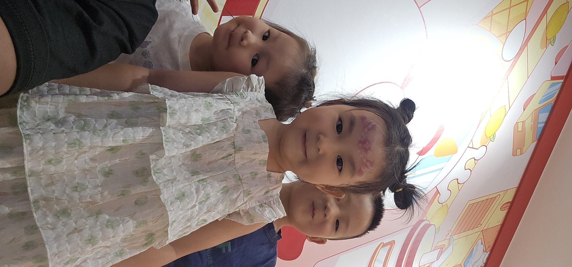 Three children, two girls and one boy, pose for a photo in front of a brightly colored wall mural. The girl in the foreground, center, has dark hair styled in two buns and a purple design painted on her forehead. She is wearing a white dress with a floral pattern. To her left, a younger girl peeks out from behind her, also with dark hair and wearing a white t-shirt. Behind the foreground girl, to the right, is a boy with short dark hair, wearing a dark blue t-shirt. The wall mural appears to depict a cartoonish scene with elements like a yellow vehicle and ice cream cones. The lighting suggests an indoor setting, possibly a public space or a party venue, given the festive mural. The image appears to be a candid snapshot taken at eye level.