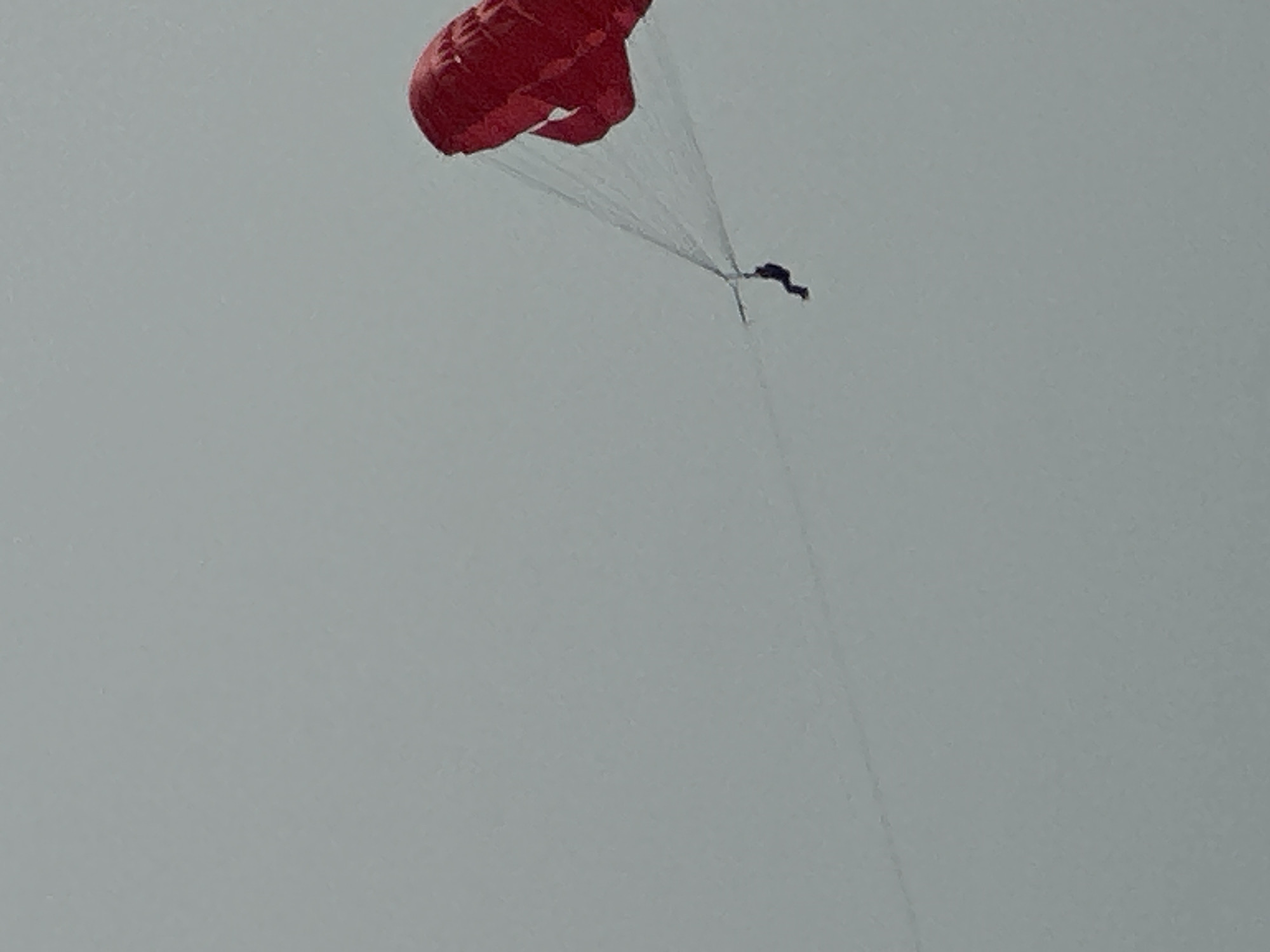 A person is parasailing against a pale gray sky. The parasail is a vibrant red, and thin white lines connect it to the person. The person is a small, dark silhouette, suspended in the air. The overall scene is simple, with no other discernible objects or structures visible. The location is not specified in the provided data.