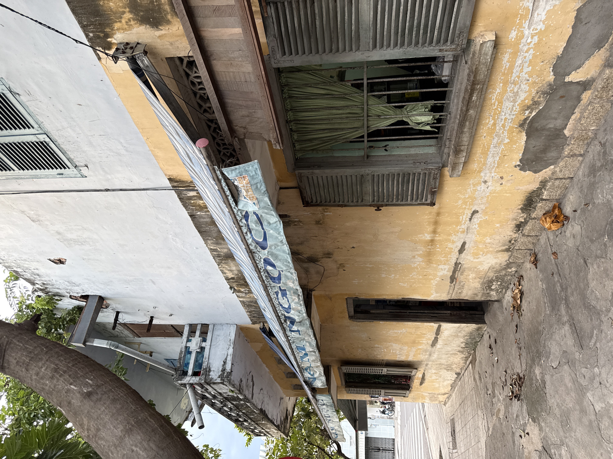 The image depicts a weathered, old building with faded yellow paint on the lower section and white paint on the upper section, located in Nha Trang, Vietnam. A large tree trunk is visible on the left, leaning over the building. The building has several features, including a balcony with a railing on the left, a sloped awning with blue lettering that appears to say "NAM NGO," two windows with wooden shutters, one of which has light green curtains, and a doorway. The ground in front of the building is a concrete sidewalk with some fallen leaves. The overall impression is one of age and disrepair, with evidence of wear and tear on the building's facade. In the background, a street with a pedestrian crossing and a few motorcycles are visible, indicating an urban setting.