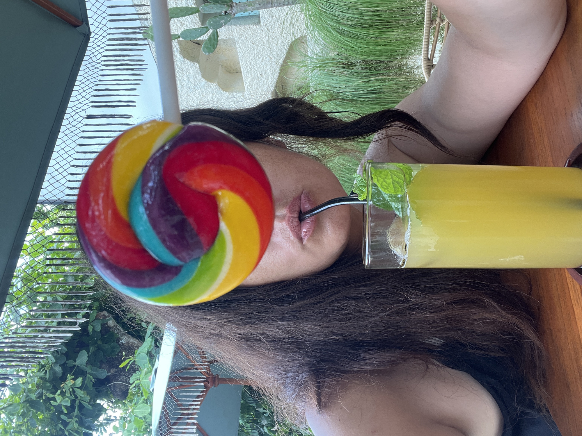 A person is seated at a wooden table with a tall glass of yellow liquid and mint leaves. They are holding a large, rainbow-colored lollipop in front of their face, obscuring it, while sipping the drink through a black straw. The background suggests an outdoor setting with lush greenery, a light-colored textured wall, and architectural elements reminiscent of Kuta Selatan, Indonesia. The lighting is natural and appears to be daytime.
