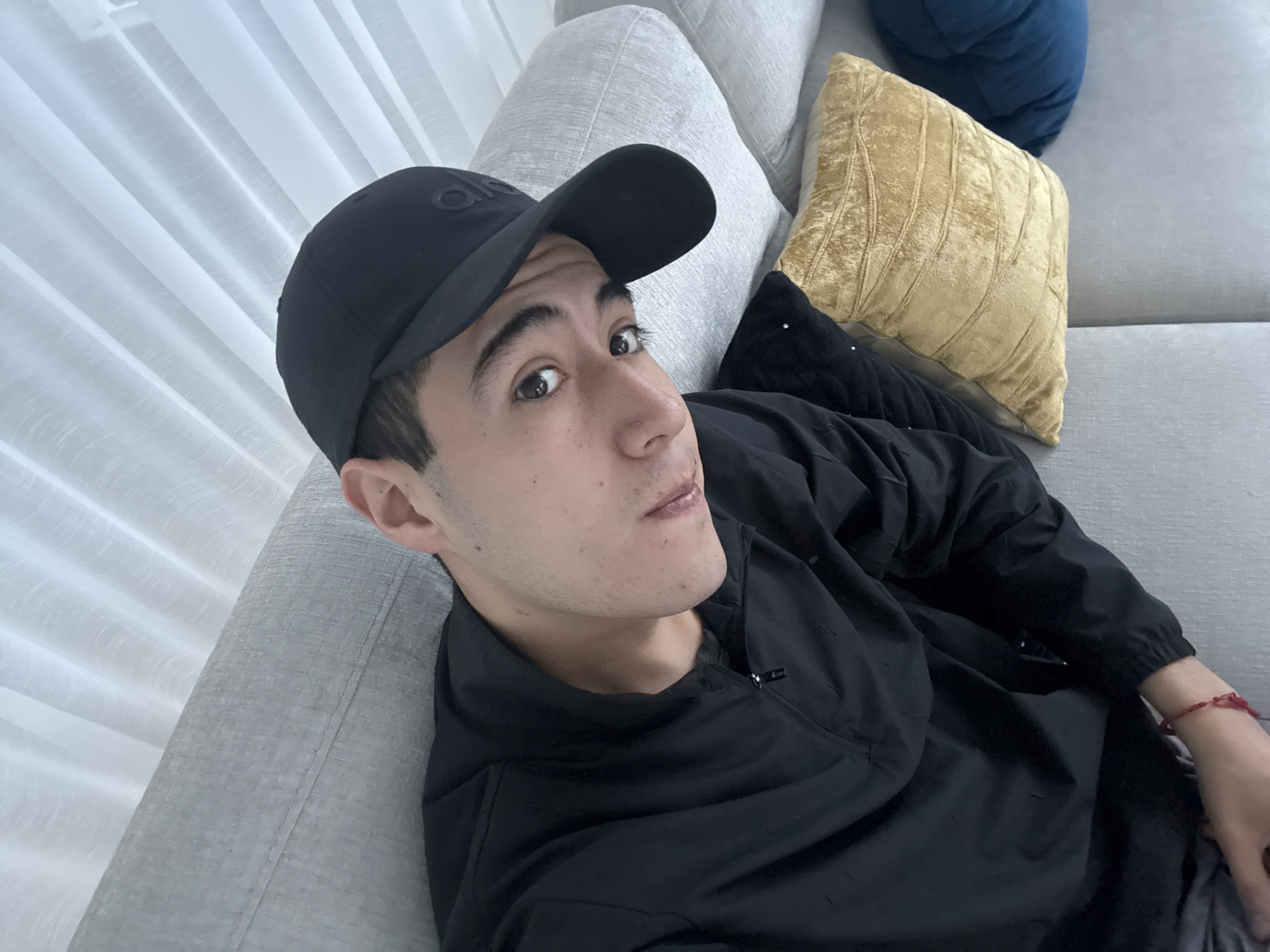 The image depicts a person lying on a light gray couch. The person is wearing a black baseball cap and a black jacket. Behind the person are several pillows: a light gray pillow, a gold-colored pillow, and a blue pillow. The setting appears to be indoors, potentially in a residence. The location is unknown.