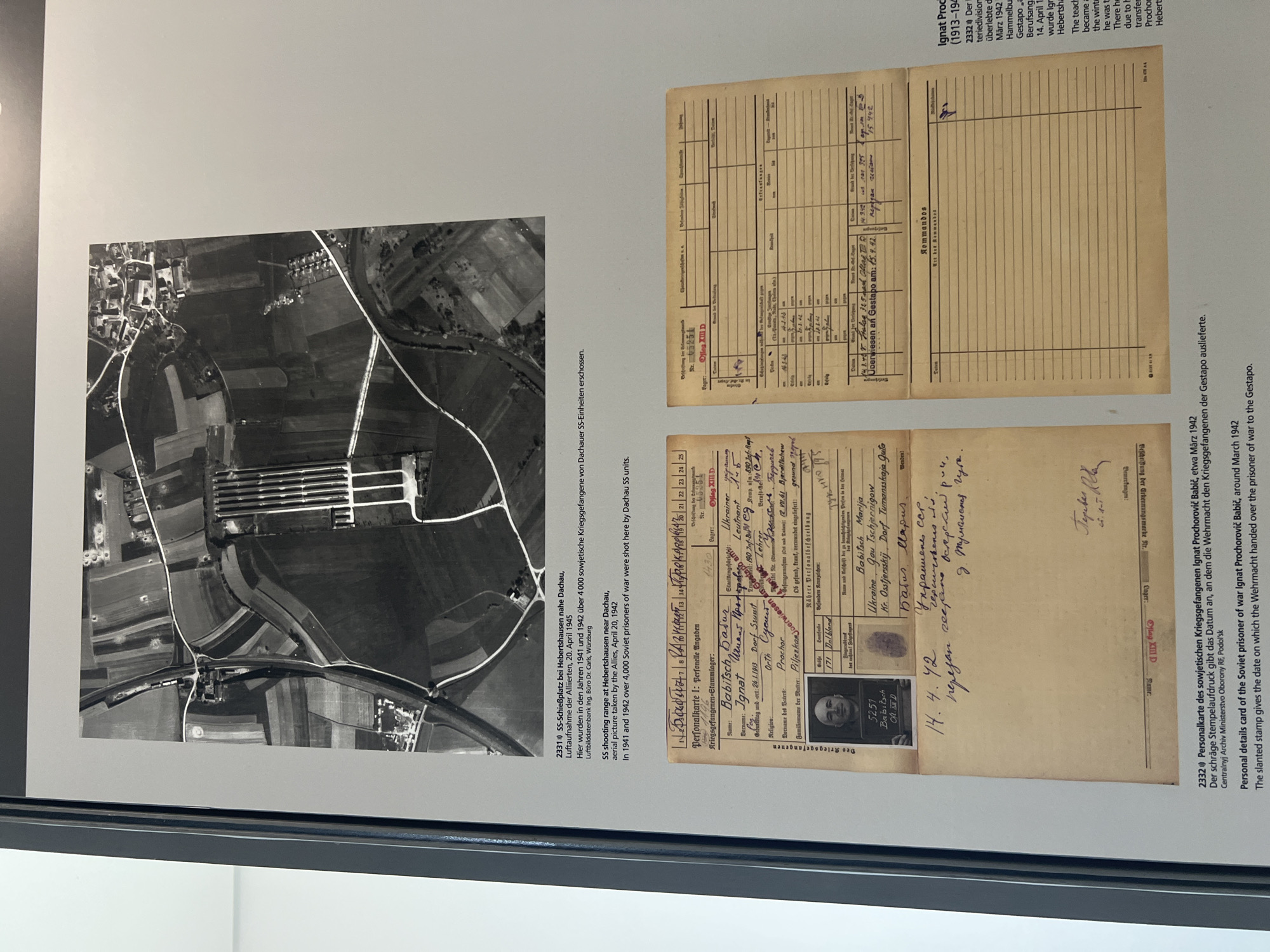 The image displays a black and white aerial photograph of a rural area with a concentration camp. The photograph is captioned with details about an SS shooting range at Hebertshausen near Dachau, Germany, and mentions that over 4,000 Soviet prisoners of war were shot there by Dachau SS units in April 1945. Below the aerial photograph is a personal detail card of a Soviet prisoner of war named Ignat Prochorovich Babič. The card, dated March 1942, contains his personal information, including his name, rank, profession, religion, and parents' names. It also includes a photograph of the prisoner and details about his transfer and release. The card is stamped and signed, and there is a handwritten note at the bottom. Another section of the card lists "Kommandos" which is partially visible. The overall scene depicts historical documentation related to World War II and the atrocities committed at the Dachau concentration camp.