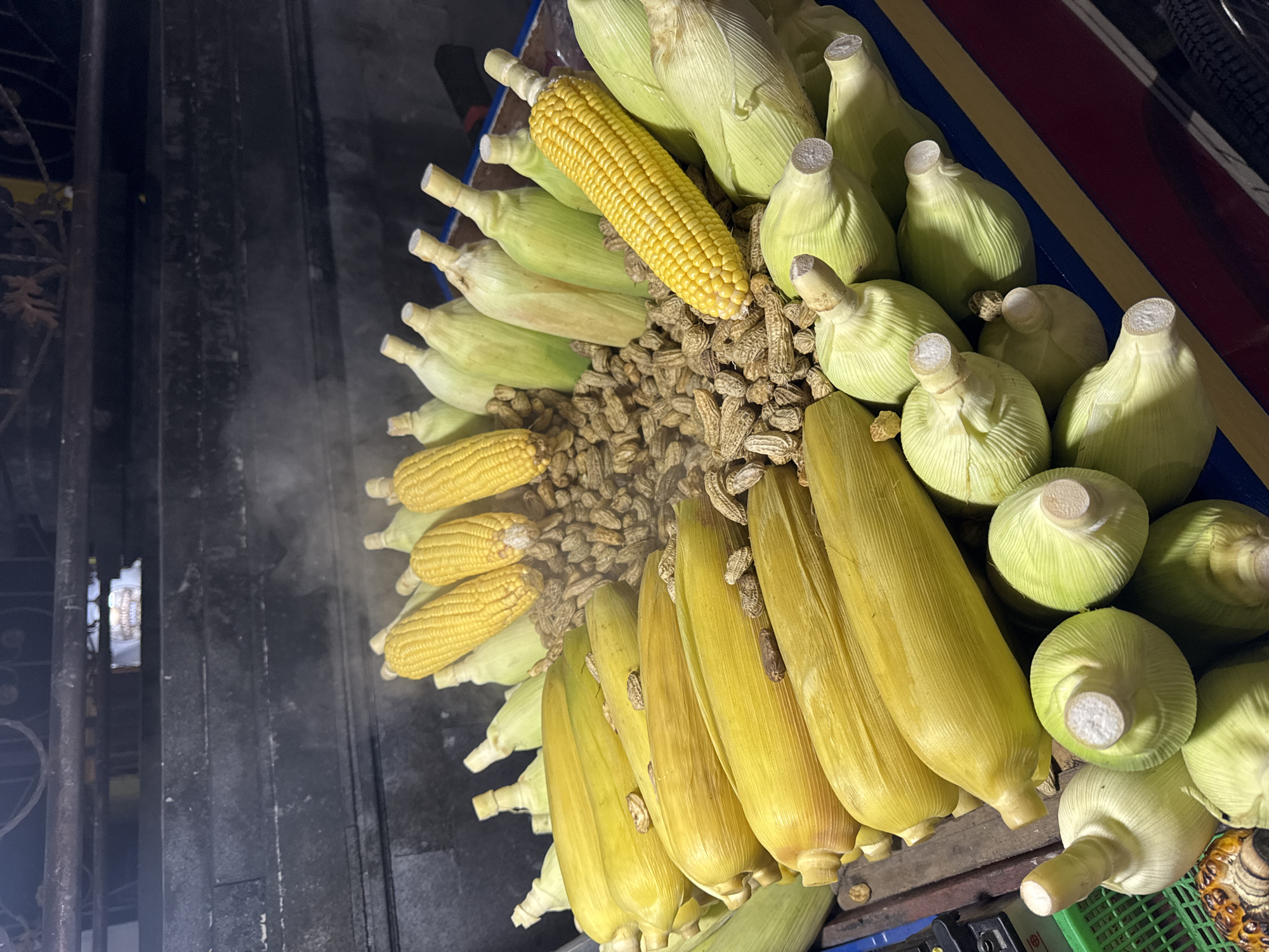 The image depicts a street food vendor in what is likely Ungasan, Indonesia, given the request's context. The vendor displays a colorful assortment of corn on the cob – some kernels exposed, others still in their green husks – and a pile of peanuts in their shells. Steam rises near the corn, suggesting it has been freshly cooked. The setup is basic, likely a cart or small table, with a dark, indeterminate background suggesting an evening or indoor setting. The scene focuses primarily on the food items ready for sale, highlighting the vendor's wares.
