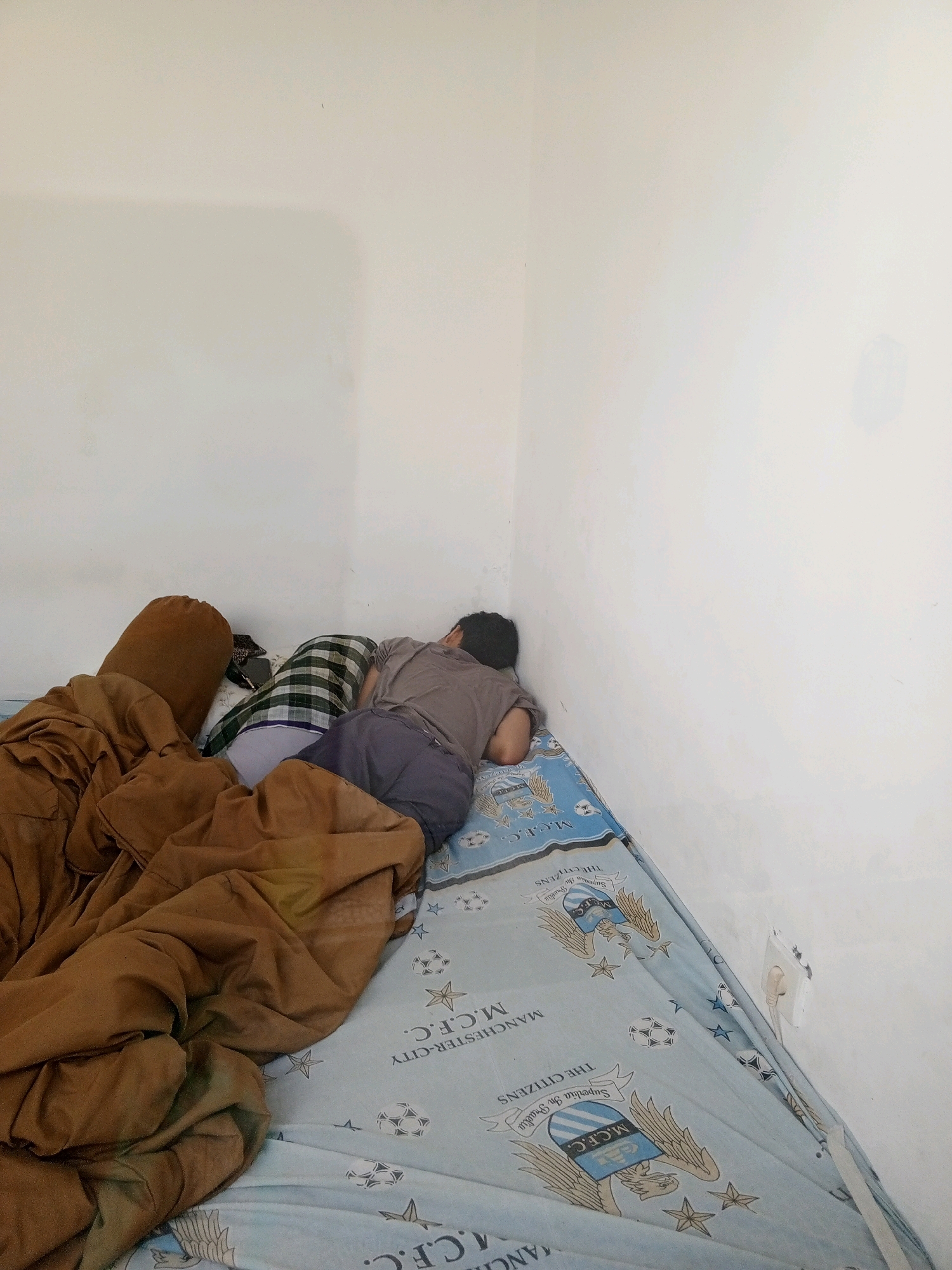 The image shows a person sleeping on a bed in a corner of a room. The walls are white and appear to be bare. The bed is covered with a light blue sheet featuring a crest and text that reads "M.C.F.C. THE CITIZENS". There are several pillows and a brown blanket on the bed. The person is lying on their side, facing the wall. An electrical outlet is visible on the right wall. The overall impression is of a simple bedroom. This scene is not specific to Denpasar, Indonesia, and could be found in any location.