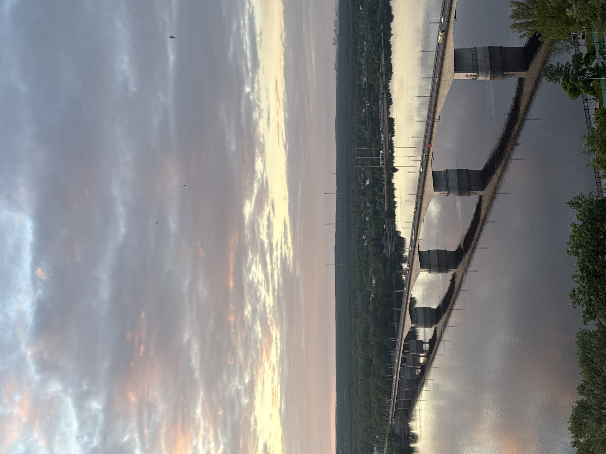 The image depicts the Kama Bridge in Perm, Russia, at dusk. The bridge, which appears to be a box girder type, stretches across the Kama River, reflecting the sky and bridge structure in the water. The sky is filled with clouds, showing hues of pink and orange, suggesting either sunset or sunrise. Dense green foliage lines the riverbanks on both sides. A road runs parallel to the river, backed by a dense forest. Distant high-rise buildings can be faintly seen on the horizon. Several birds are visible in the sky. Cars are seen driving on the bridge.