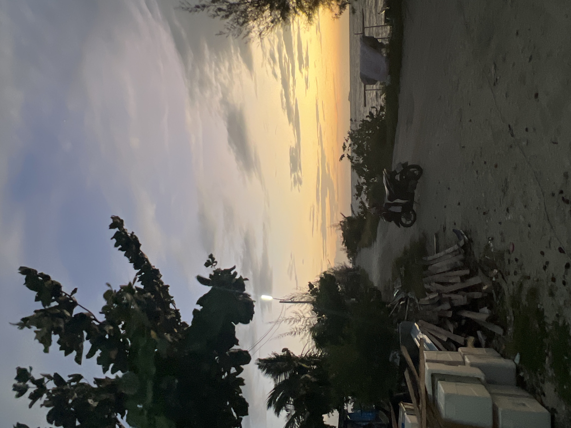 The image depicts a dirt road leading towards a body of water during a sunset. A scooter is parked on the right side of the road. To the left of the road, there are stacks of what appear to be building materials, including white rectangular foam blocks and wooden planks. Trees and vegetation are present on both sides of the road and in the foreground. The sky is filled with clouds illuminated by the setting sun, casting orange and yellow hues. A street lamp is visible on the left side of the road, and wires extend from it. The location is described as being in the Ko Pha-ngan District of Thailand.