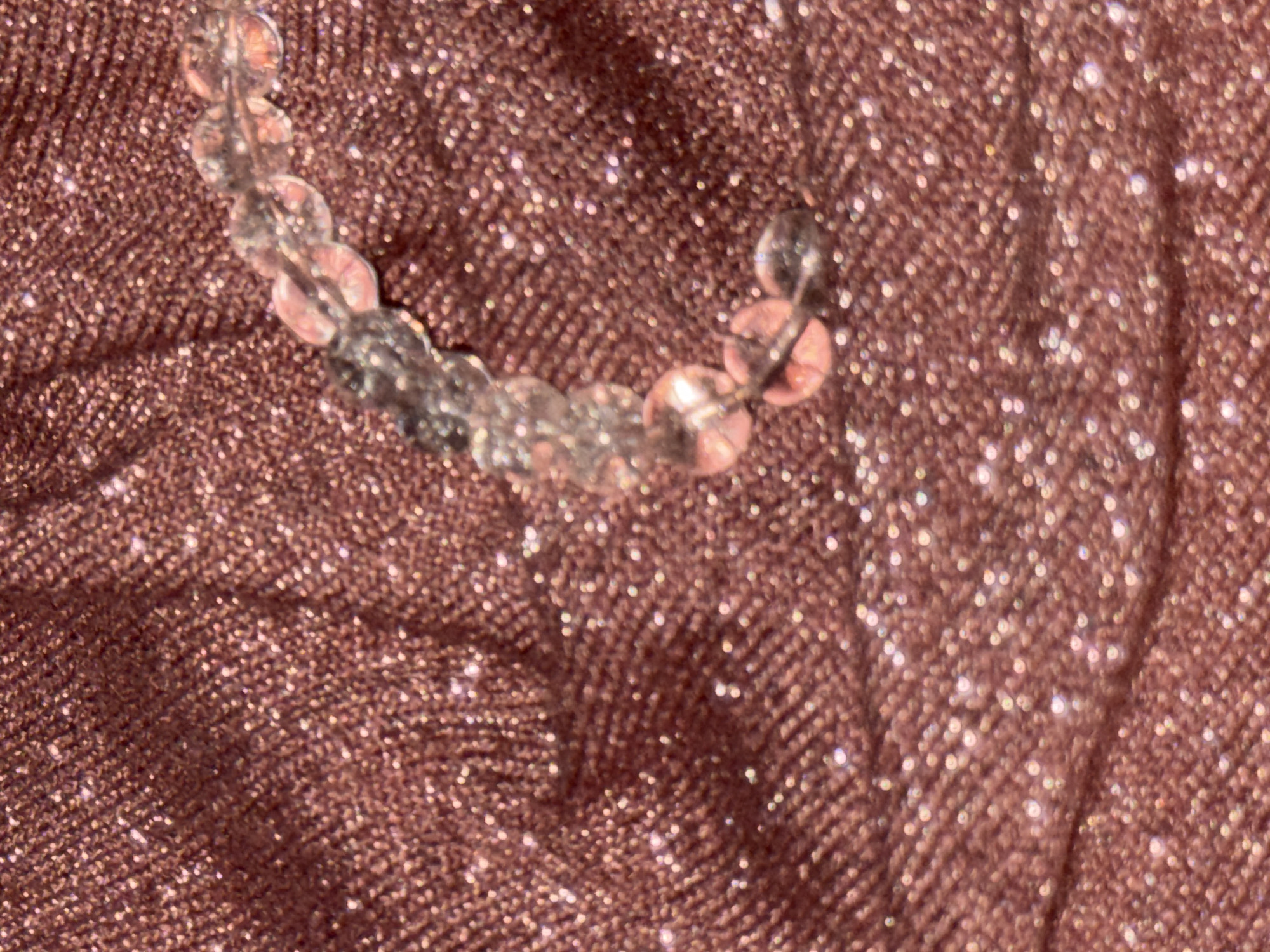 A close-up shot shows a semi-transparent beaded bracelet resting on a shimmering, reddish-brown fabric. The beads have a translucent appearance with faint pinkish hues. The background fabric has a textured, glittering surface. The location in the city of Chaikovsky, Russia is not evident from the image.