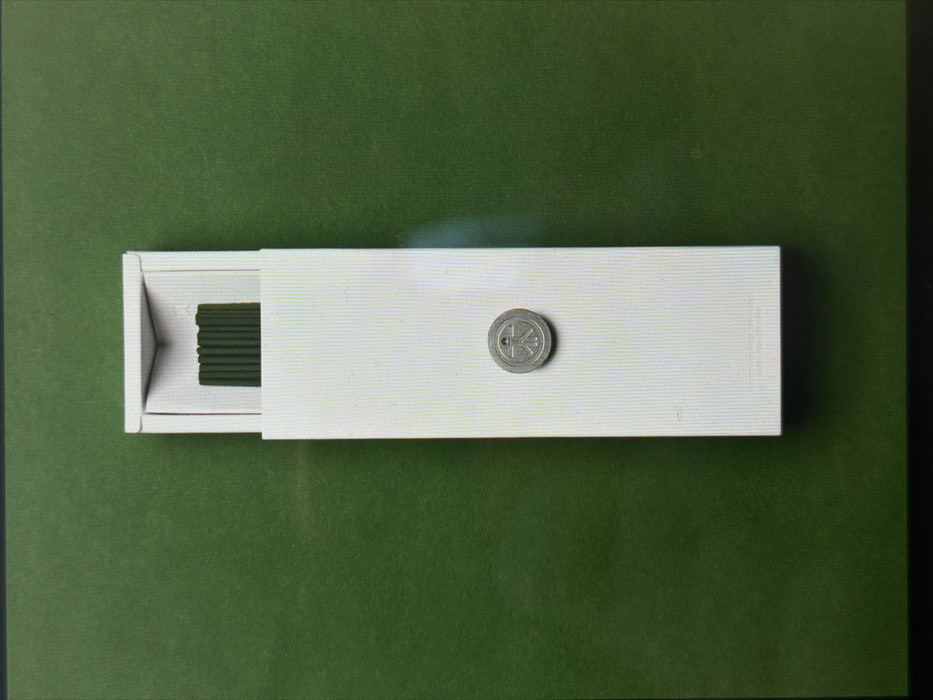 A vertically oriented, elongated white rectangular box is centered against a uniform, solid dark green background. The top flap of the box is partially open, revealing a neat row of approximately nine thin, dark green cylindrical objects inside. A circular, metallic, silver-colored disc is positioned centrally on the front surface of the white box, approximately two-thirds of the way down from the top. The disc features a clearly embossed or engraved symbol depicting a stylized human figure with outstretched limbs contained within a circular outline. No individuals or active interactions are depicted. The scene is static, illuminated evenly, with a minor glare visible on the right side of the white box. Location in the city of London, United Kingdom, cannot be determined from the visual evidence provided.