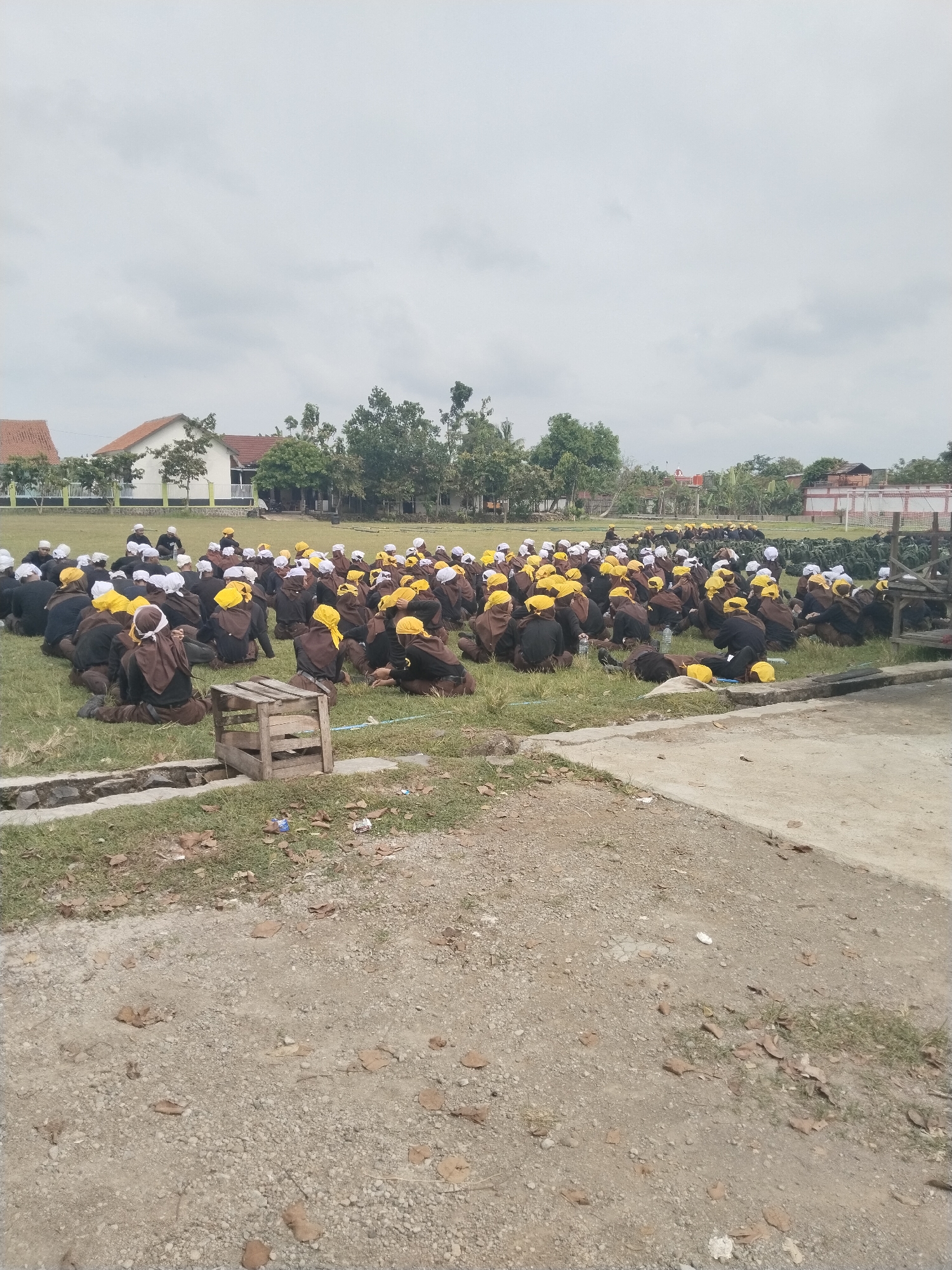 A large group of individuals, predominantly young people, are seated on a grassy field under an overcast sky during daytime. Most are wearing dark brown or black uniforms or similar attire, complemented by brightly colored head coverings โ a majority in vivid yellow, with some wearing white. They are arranged in rows, facing generally towards the right side of the frame, suggesting they are attending an organized event or assembly, possibly a training session or a school gathering in Tawangsari, Indonesia.
In the foreground, a rough dirt and gravel path with scattered dry leaves occupies the lower portion of the image. A concrete drain or channel separates this path from the grassy area where the people are seated. A rustic wooden crate or frame is prominently placed near the drain, close to the seated group on the left.
The background features several houses with reddish-orange tiled roofs and white walls, partially obscured by various trees and lush greenery. A white and green fence runs behind some of the houses. Further to the right in the background, stacks of what appear to be military or outdoor training gear, possibly green camouflage bags or equipment, are visible. The overall setting suggests an outdoor institutional or communal ground in a semi-urban or rural area. The light is diffused, indicating a cloudy or overcast day. No discernible text is visible in the image.